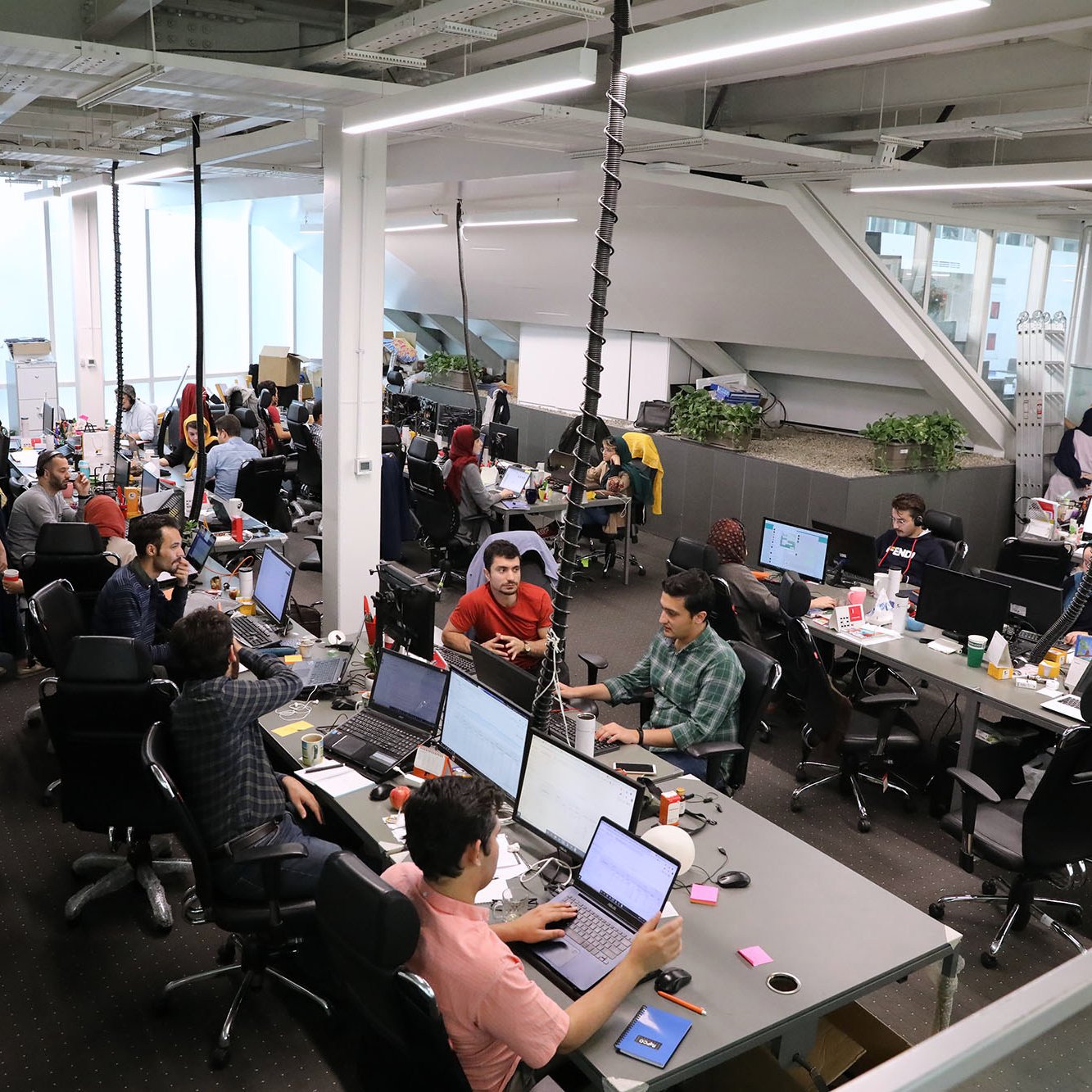 L'image montre un espace de bureau moderne et dynamique. Il y a plusieurs personnes qui travaillent à des bureaux équipés d'ordinateurs. L'environnement semble collaboratif, avec des employés assis autour de tables, certains en train de discuter, tandis que d'autres se concentrent sur leur travail. Les bureaux sont bien éclairés et décorés avec des plantes. On peut voir des objets personnels et des collations éparpillés sur les tables, ce qui donne une atmosphère informelle et conviviale.