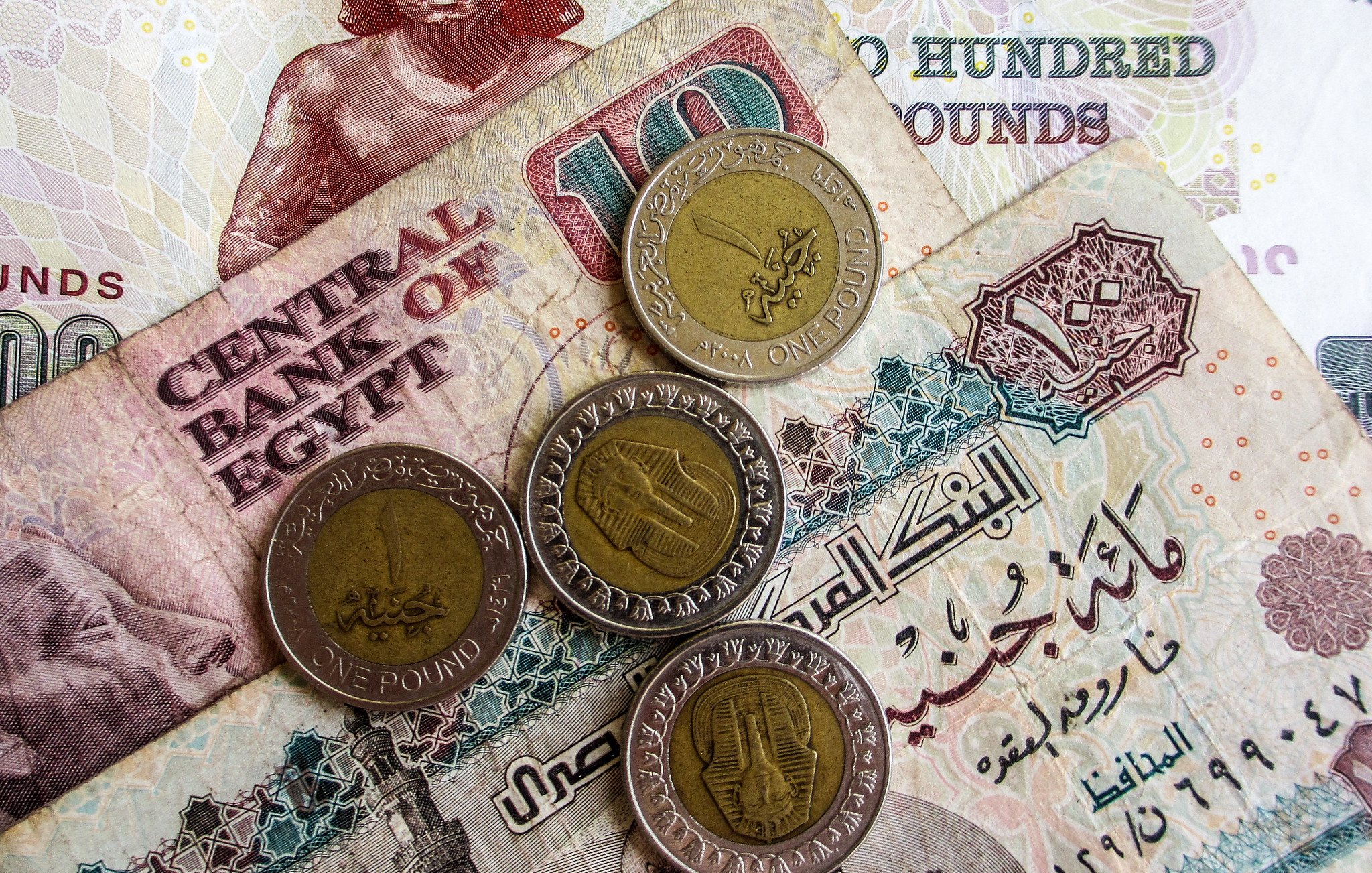 L'immagine mostra diverse banconote e monete egiziane. In primo piano ci sono quattro monete, due delle quali sono di un pound egiziano. Sullo sfondo, ci sono banconote di diverse denominazioni, evidentemente con scritte arabe, tipiche della valuta egiziana. Una banconota sembra avere il valore di cento pounds e presenta un motivo decorativo. L'atmosfera è quella di un contesto economico, evidenziando elementi tipici della valuta dell'Egitto.