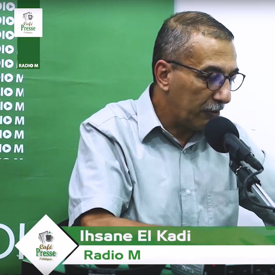 L'image montre un homme assis derrière un micro dans un studio de radio. Il porte une chemise à manches courtes et des lunettes. Sur la table devant lui, il y a un verre d'eau et un téléphone. L'arrière-plan est vert avec des éléments de branding pour "Radio M". Le nom "Ihsane El Kadi" est affiché en bas de l'image.