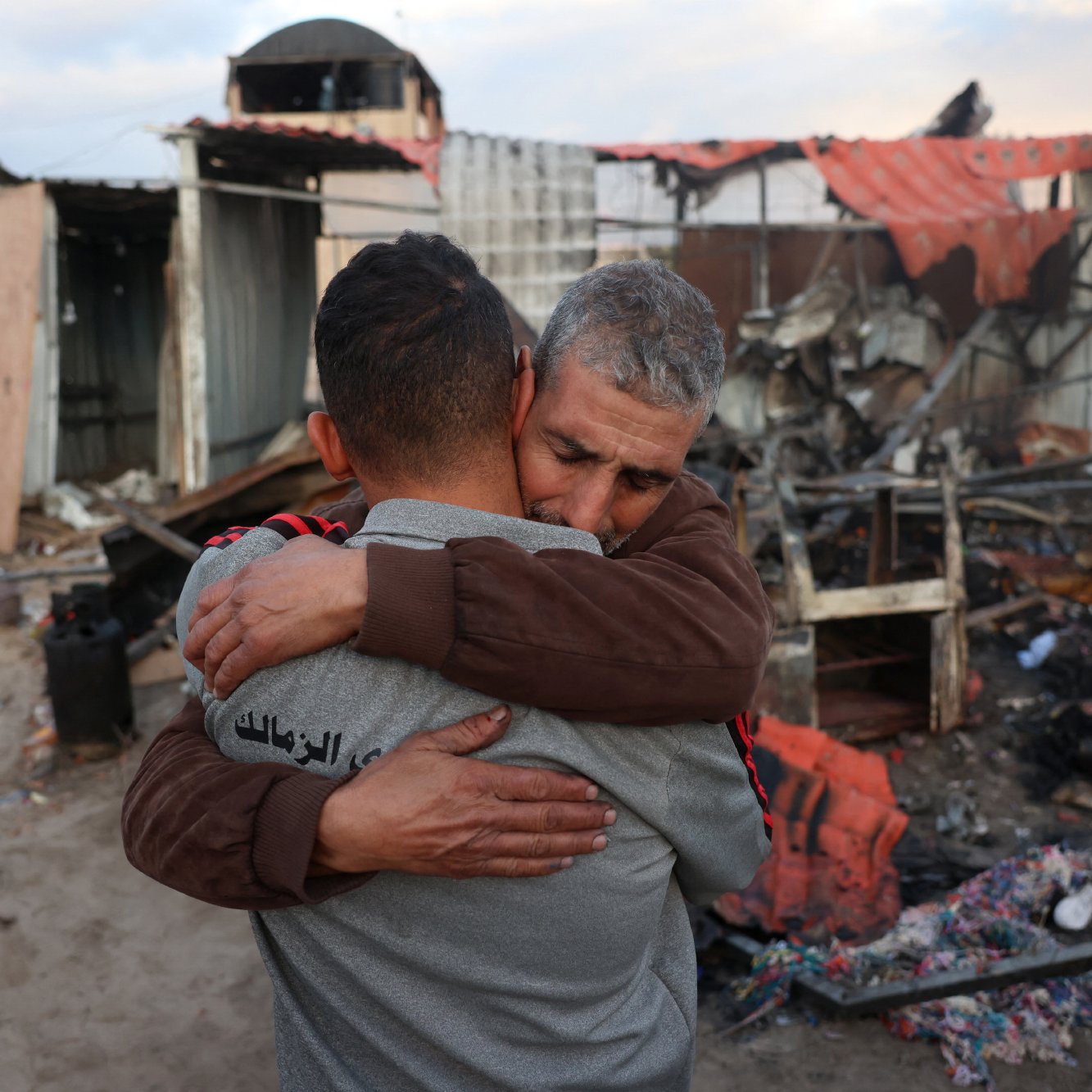 L'image montre deux hommes qui s'étreignent de manière émotive au milieu de décombres. En arrière-plan, il y a des restes de bâtiments détruits, avec des morceaux de métal et des débris éparpillés. L'ambiance semble très chargée d'émotion, suggérant une perte ou un chagrin. Les expressions des hommes témoignent d'une connexion forte et d'un besoin de réconfort dans un moment difficile.