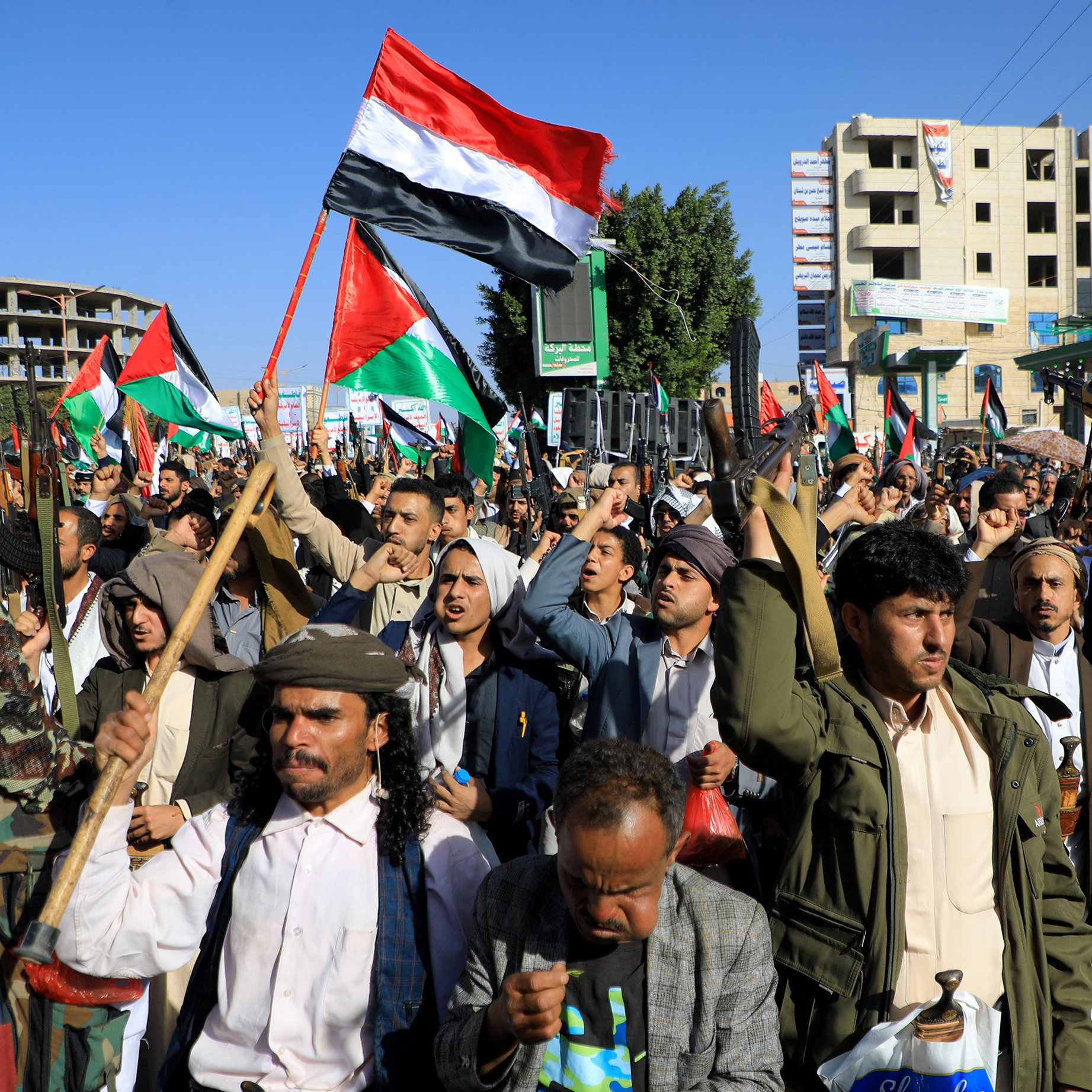 L'immagine mostra una folla di persone in protesta, che si riunisce in una manifestazione. I manifestanti alzano bandiere, probabilmente di carattere nazionale o politico, e sembrano esprimere il loro fervore con entusiasmo. Molti indossano abiti tradizionali e ci sono anche elementi di armi, come fucili, che suggeriscono una manifestazione di forte impatto emotivo e potenzialmente conflittuale. Gli edifici sullo sfondo indicano un contesto urbano, mentre l'atmosfera complessiva sembra intensa e carica di significato.