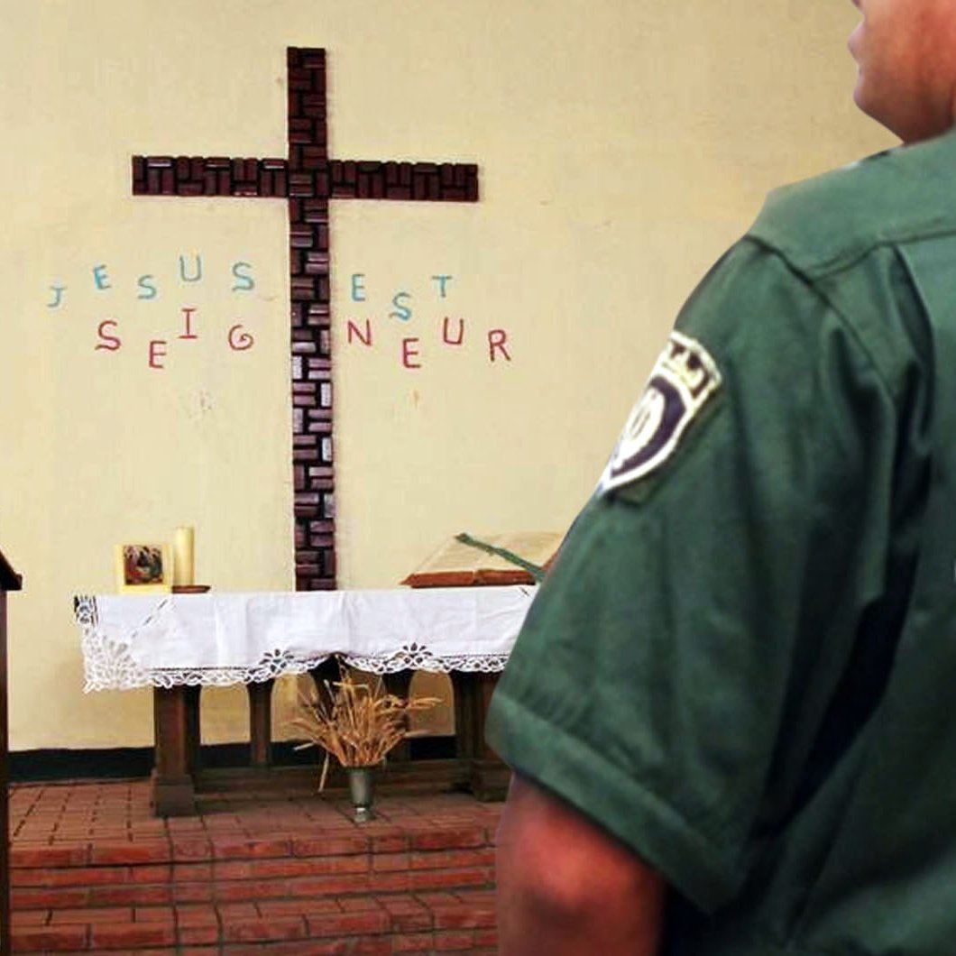 L'image montre un intérieur d'une église avec une grande croix en bois sur le mur. Des mots sont écrits autour de la croix, où l'on peut lire "JÉSUS EST SEIGNEUR". Sur la gauche, il y a une table d'autel avec un drapé. À droite, un homme en uniforme vert, probablement un agent de police ou de sécurité, est vu de dos, portant un badge qui indique "الدرك الوطني" en arabe, ce qui signifie "Gendarmerie nationale". L'ambiance de l'image semble calme et spirituelle, mêlant des éléments religieux et de sécurité.