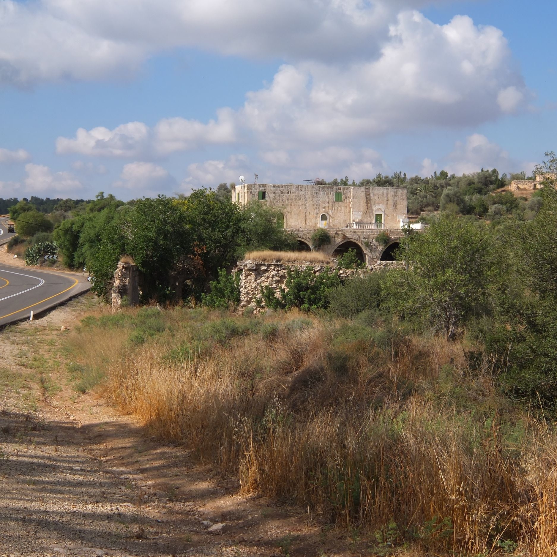 L'immagine mostra un paesaggio rurale caratterizzato da una strada curvilinea che si snoda tra la vegetazione. Ai lati della strada, ci sono prati e aree erbose, con un'atmosfera tranquilla e naturale. Sullo sfondo, spicca una vecchia costruzione in pietra, apparentemente in rovina, con archi visibili, circondata da alberi e vegetazione. Nel cielo, si possono vedere alcune nuvole sparse, che completano il quadro di un ambiente sereno e suggestivo.