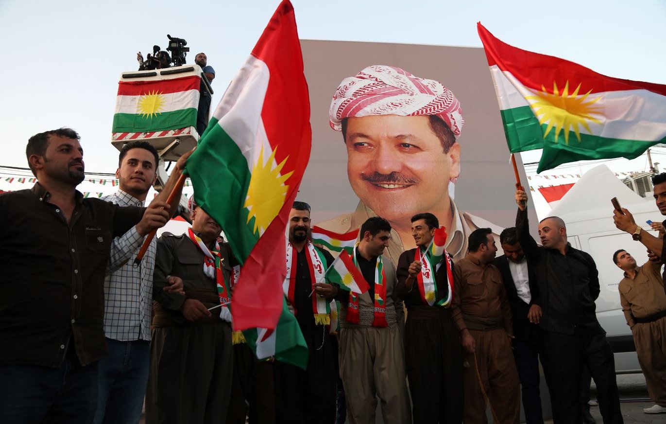 L'image montre un groupe de personnes tenant des drapeaux kurdes, célébrant devant une grande affiche d'une personnalité kurde. Les participants portent des vêtements traditionnels et semblent joyeux, suggérant une ambiance festive. L'affiche derrière eux représente un homme souriant, probablement un leader ou une figure importante dans la culture kurde. Les décorations et les drapeaux créent une atmosphère vibrante et patriotique.