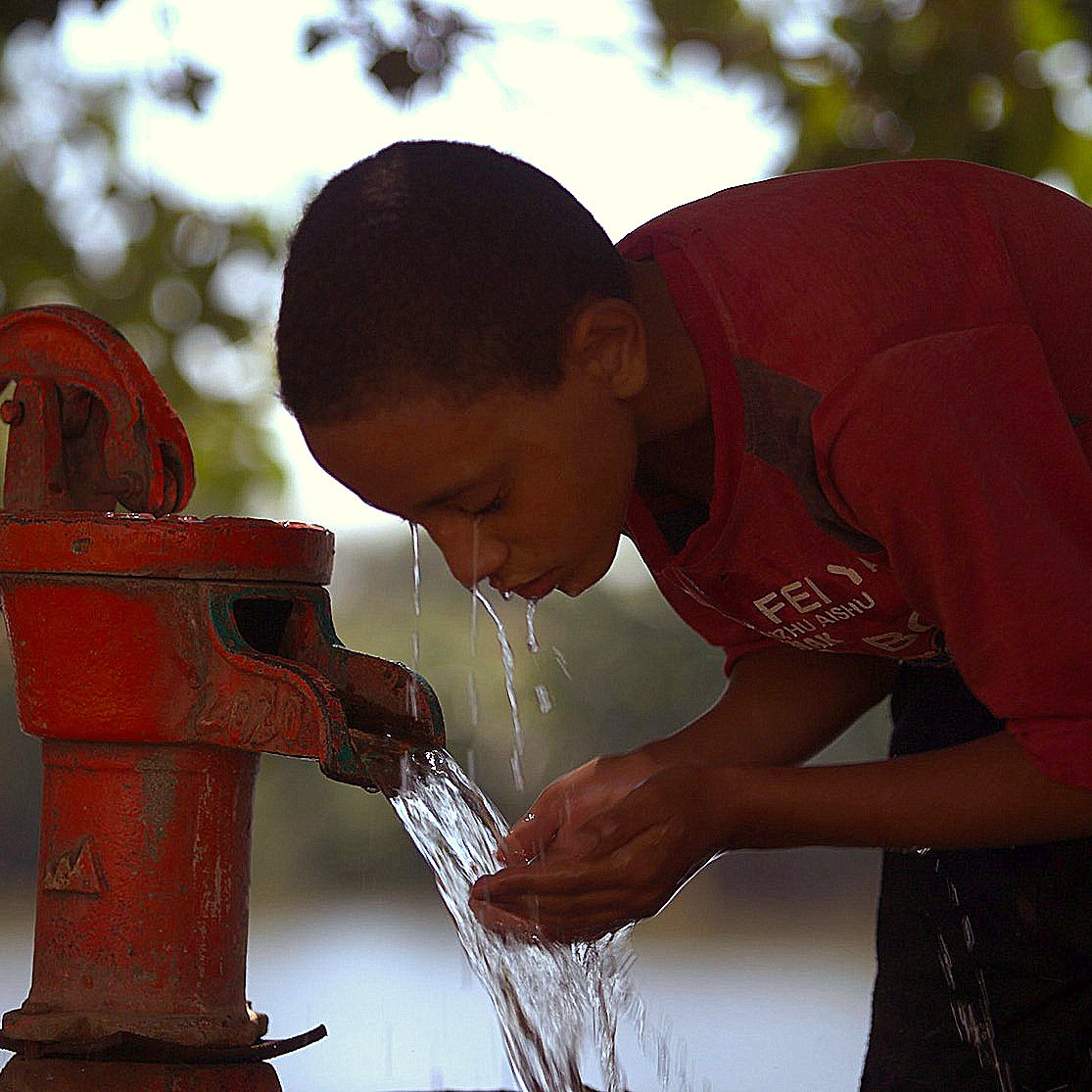 The image shows a young boy bent over a red hand pump, drawing water from it. He appears focused as he cups his hands to catch the water flowing out. The background features greenery and suggests a natural setting, possibly near a body of water, with soft light creating a warm atmosphere. The boy is wearing a red shirt, and the scene conveys a sense of simplicity and the importance of access to fresh water.