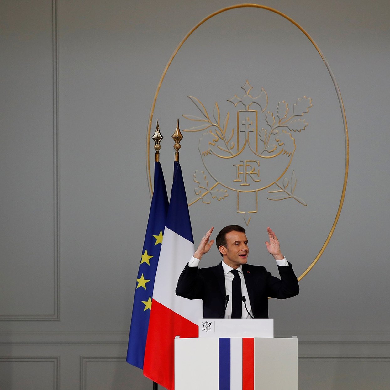 در این تصویر، یک مرد در حال سخنرانی است. او در جلوی یک تریبون ایستاده و دست‌هایش را به سمت بالا برده است. در پشت او، نماد رسمی فرانسه وجود دارد که شامل دو پرچم می‌باشد؛ یکی پرچم فرانسه و دیگری پرچم اروپایی. دیوار پشت او ساده و به رنگ خاکستری است و نماد جمهوری فرانسه در مرکز قرار دارد. این صحنه نشان‌دهنده یک رویداد رسمی یا کنفرانس مطبوعاتی است.
