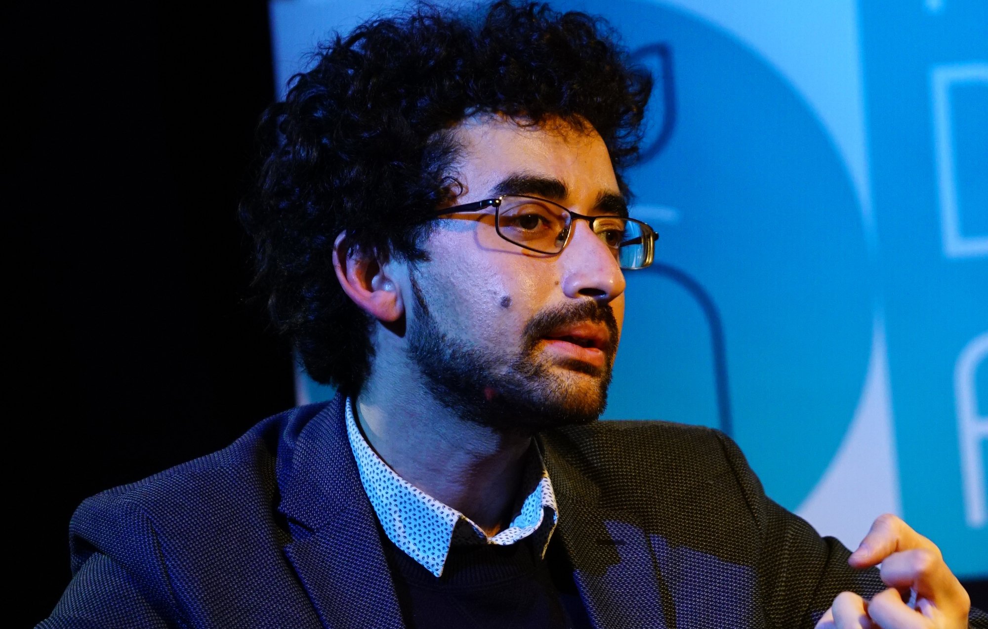 Homme avec des lunettes, cheveux bouclés, en discussion, vêtu d'une veste.