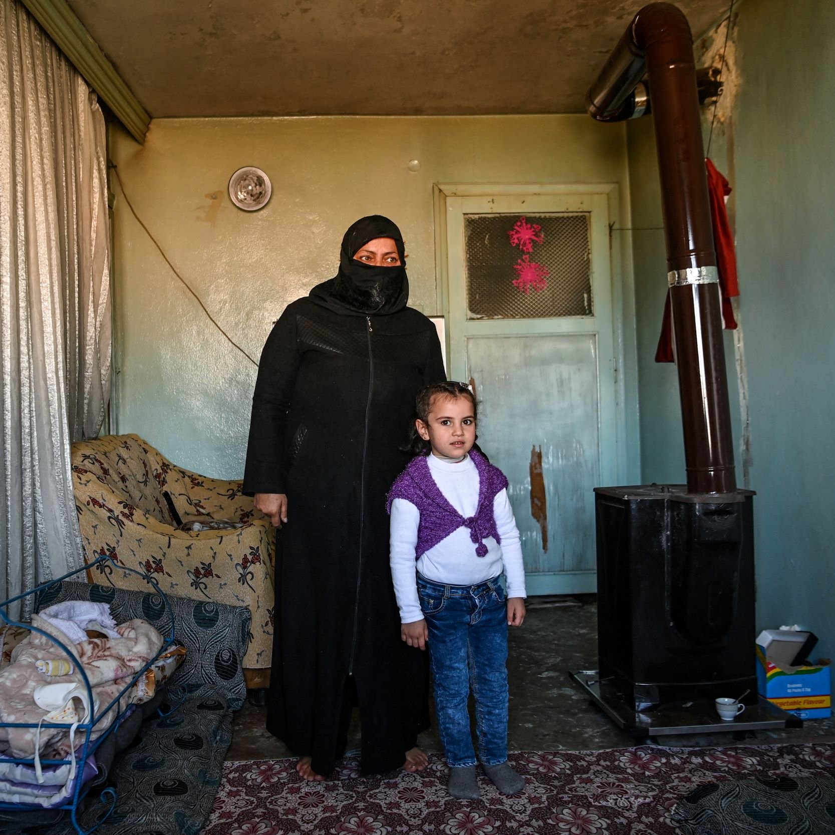 L'image montre une femme portant une tenue noire et un voile, se tenant à côté d'une petite fille. Elles sont à l'intérieur d'une pièce qui semble modeste, avec des murs verts pâles et un tapis au sol. À gauche, il y a une couchette avec un bébé endormi. Un poêle en métal est visible dans un coin de la pièce, et des rideaux en tissu léger sont tirés. L'ambiance est familiale, mais la simplicité du décor reflète une certaine précarité.