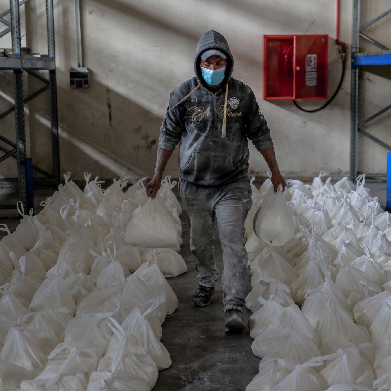L'immagine mostra un uomo che cammina in un ambiente di magazzino. Indossa una felpa con cappuccio e una mascherina e tiene in mano due sacchetti bianchi. Intorno a lui ci sono numerosi sacchetti bianchi disposti ordinatamente sul pavimento. Le pareti del magazzino sono spoglie e ci sono scaffali metallici sullo sfondo. L'atmosfera sembra lavorativa e organizzata.