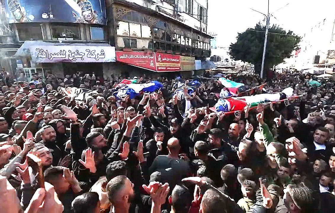 L'image montre une grande foule rassemblée dans une rue, avec des personnes portant des drapeaux et des pancartes. Il y a des cercueils sur des supports, symbolisant un hommage ou des funérailles. La foule est visiblement très engagée, levant les mains et exprimant des émotions fortes. L'environnement urbain est marqué par des bâtiments et des affiches en arrière-plan. L'atmosphère est chargée de gravité et de solidarité.