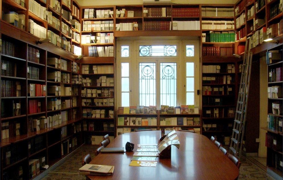 L'image montre une bibliothèque élégante et bien organisée. Les étagères sont remplies de livres sur plusieurs niveaux, allant du sol au plafond. Au centre, il y a une grande table en bois entourée de chaises, créant un espace propice à la lecture ou à l'étude. La lumière naturelle pénètre par de grandes fenêtres avec des motifs décoratifs, apportant une ambiance chaleureuse et accueillante. L'ensemble de l'endroit évoque un sentiment de calme et de sérénité, idéal pour les passionnés de lecture.