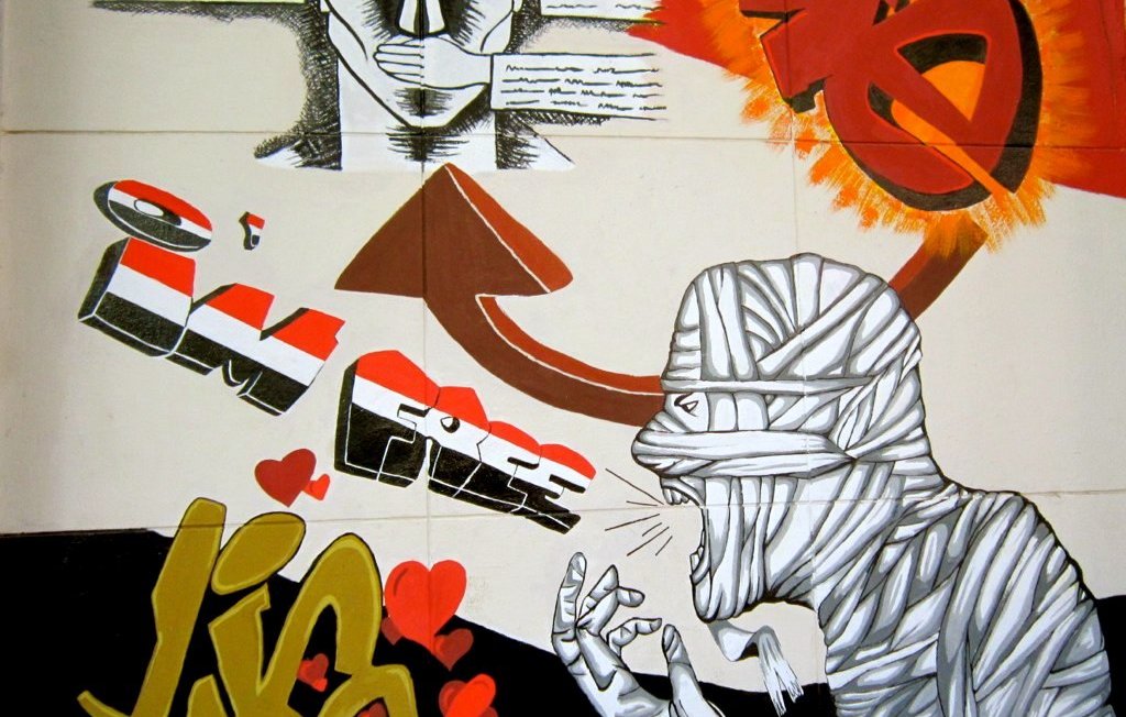 L'image représente une œuvre de street art colorée. On y voit un personnage avec des lunettes de soleil et une expression déterminée, entouré de textes. Au-dessus, une flèche pointe vers le haut avec l'inscription "I'M FREE" (je suis libre) en lettres stylisées. À droite, un autre personnage est représenté, désigné comme étant bandé, avec des gestes qui semblent exprimer une forte émotion. Le fond combine des couleurs vives comme le rouge et le jaune, avec des cœurs dessinés au bas, ce qui ajoute une touche plus douce à l'ensemble. L'œuvre montre un contraste entre la liberté et une certaine souffrance, évoquant des thèmes profonds.