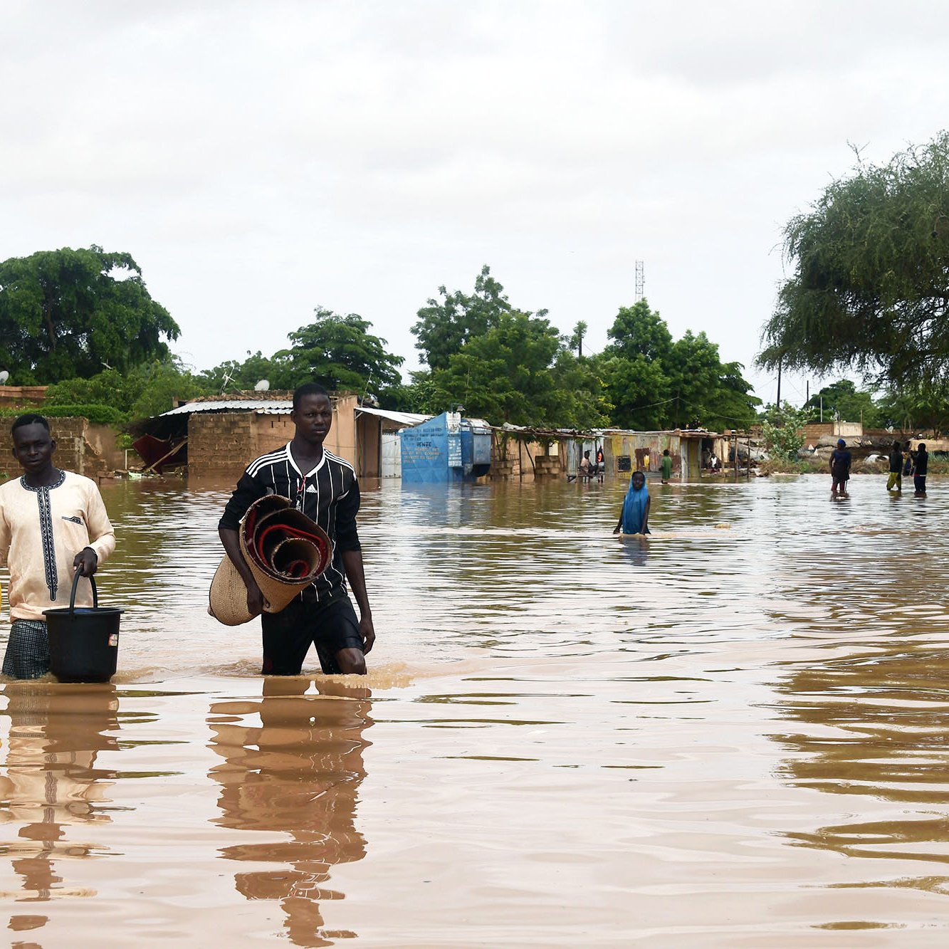 The image depicts a flooded area, where individuals are walking through standing water. Two men are central in the foreground; one is carrying a bucket, while the other holds a rolled-up mat. In the background, makeshift shelters or homes are partially submerged, surrounded by trees and greenery. The scene reflects the impact of flooding, with water covering the ground and affecting the surrounding environment. The sky is overcast, suggesting ongoing adverse weather conditions.