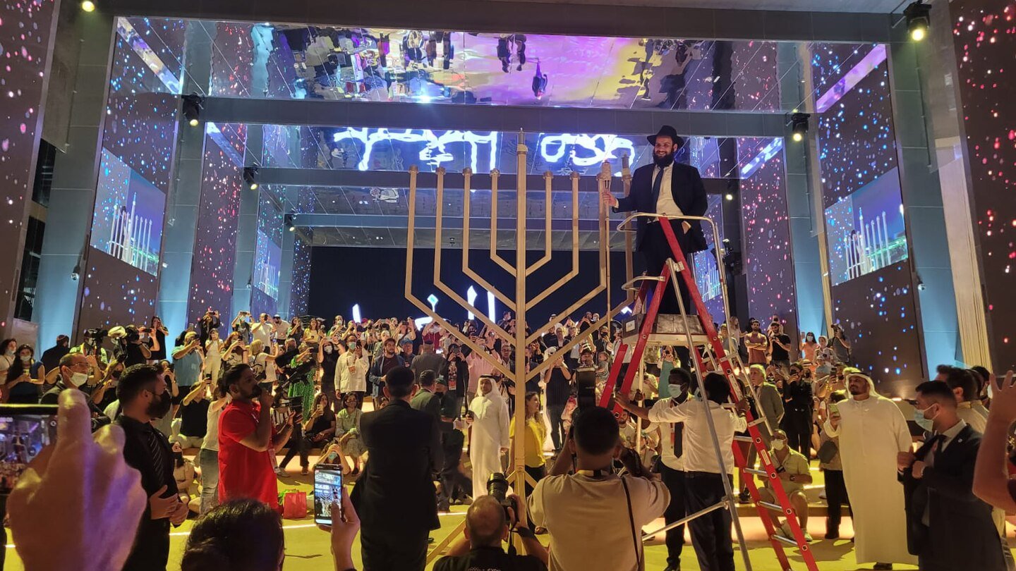 الصورة تظهر حدثًا احتفاليًا في مكان كبير. في المركز، يوجد هيكل على شكل منارة (المناورة)، مما يشير إلى الاحتفال بمناسبة معينة. يتم تنظيم الحضور حول الهيكل، وهناك أشخاص يحملون هواتفهم لتصوير الحدث. بعض الحضور يرتدون ملابس تقليدية، بينما يرتدي آخرون ملابس رسمية. هناك إضاءة ملونة وتفاصيل زينة جميلة في الأجواء، مما يضيف جوًا احتفاليًا حيويًا.