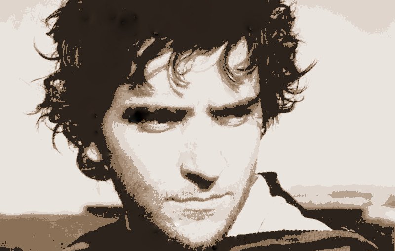 L'image montre un homme avec des cheveux bouclés et une expression pensive. Il porte un pull à rayures et regarde au loin, ce qui laisse transparaître une certaine intensité dans son regard. Le fond est flou, ce qui met davantage l'accent sur son visage. Les couleurs semblent délavées, donnant une impression nostalgique à l'ensemble de la scène.