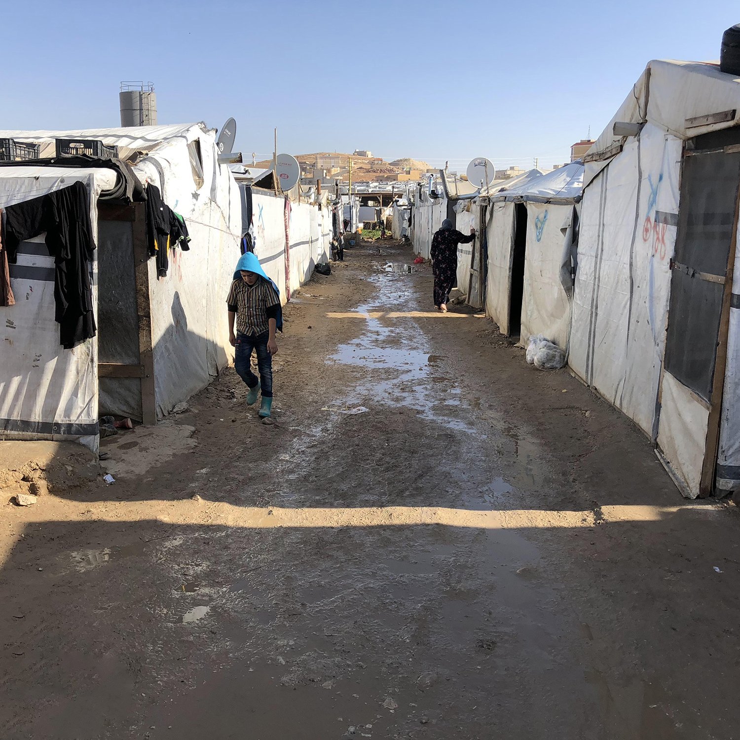 The image depicts a narrow path between rows of temporary tents or makeshift shelters, likely in a refugee or displaced persons camp. The tents are primarily white and appear to be made from tarpaulin or similar materials. There are clothes hanging outside some of the tents, suggesting attempts to dry them. The ground is muddy, indicating recent rain, and the surrounding area appears arid with few visible structures beyond the camp. The scene conveys a sense of modest living conditions and the challenges faced by individuals in such environments.