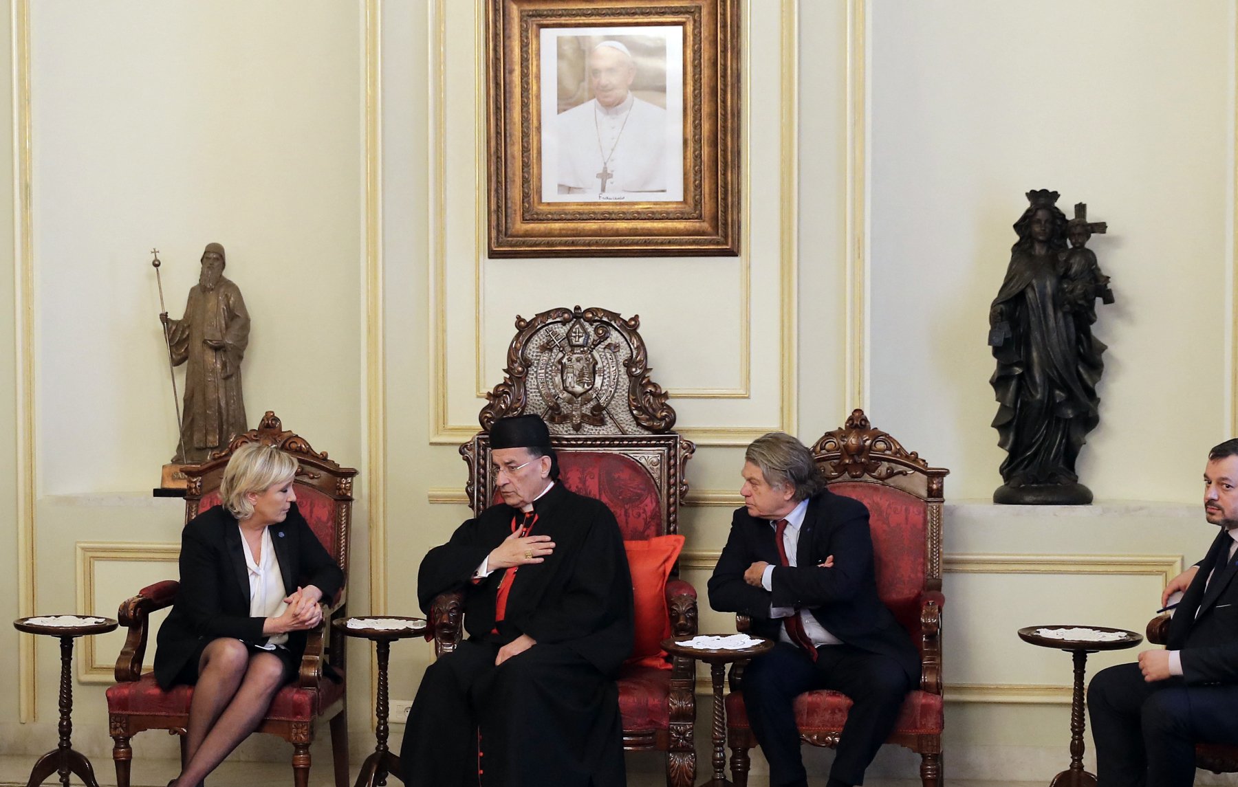 در این تصویر، یک جلسه‌ای در یک اتاق رسمی دیده می‌شود. در مرکز تصویر، یک مرد با لباس مذهبی نشسته است که ظاهراً یک مقام دینی است. او دستش را بر روی قلبش قرار داده و به دو نفر دیگر که در طرفین او نشسته‌اند، توجه می‌کند. زن سمت چپ تصویر گویی در حال گوش دادن به صحبت‌های اوست و مرد سمت راست تصویر نیز در حال بحث یا تبادل نظر به نظر می‌رسد. محیط اتاق تزئین شده و شامل یک تابلو در دیوار و مجسمه‌هایی در پس‌زمینه است. فرش زیر پای آنها نیز به فضای رسمی‌ و سنتی اتاق افزوده است.