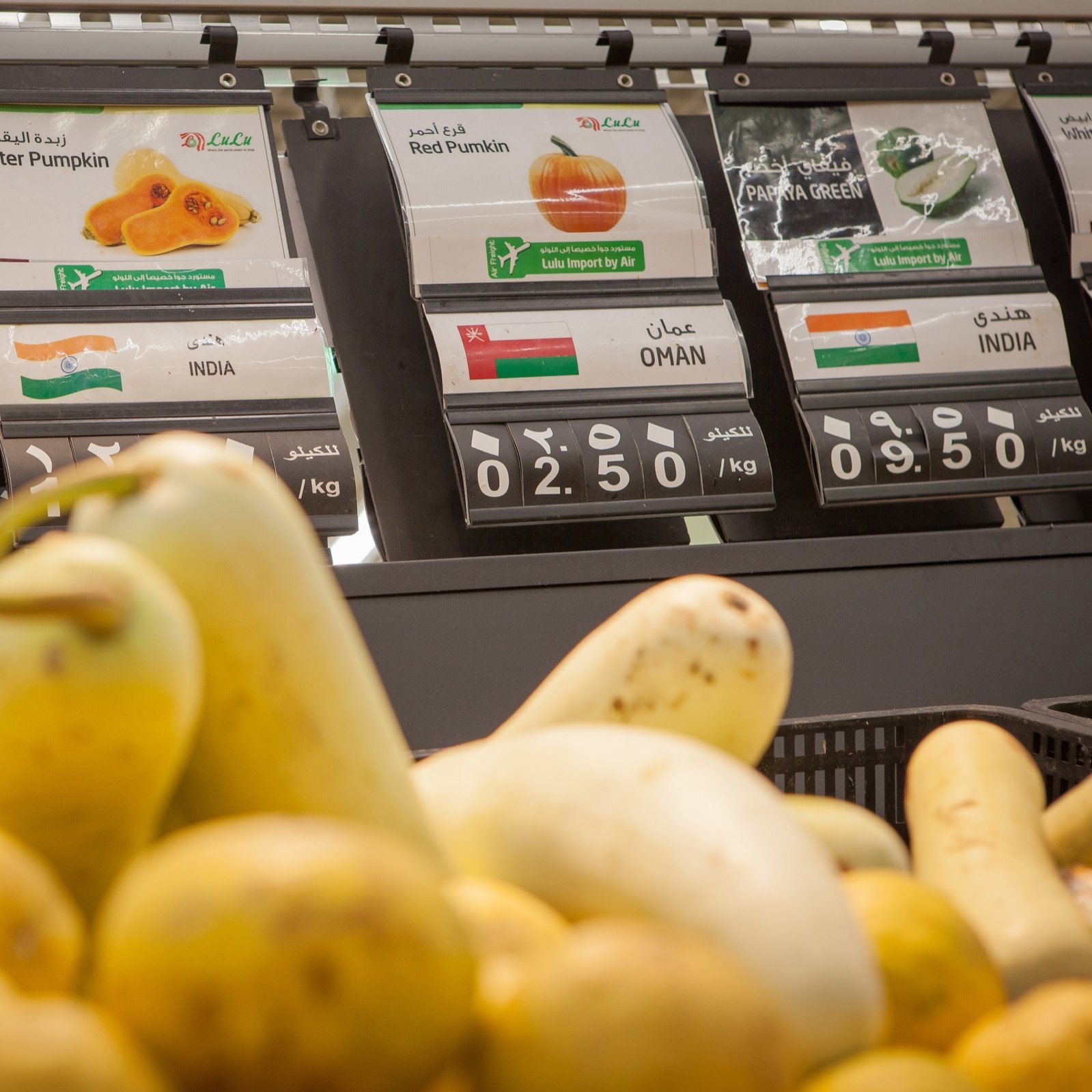 L'image présente un banc de marché dans un magasin, où divers types de courges sont exposés. Au premier plan, on voit des courges de différentes formes et couleurs. En arrière-plan, des étiquettes indiquent les prix et les origines des produits, avec des mentions comme "Courge blanche" et "Courge butternut", ainsi que des pays d'origine tels que l'Iran, Oman et l'Inde. Les prix sont affichés en kilos. L'ensemble crée une ambiance de supermarché frais et coloré.