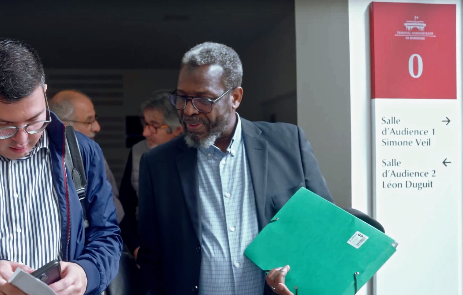 L'image montre un groupe de personnes dans un couloir apparemment nimbé d'une ambiance professionnelle. Deux hommes au premier plan interagissent, l'un tenant un téléphone et l'autre un dossier vert. L'environnement semble être celui d'un bâtiment public ou d'un tribunal, avec des panneaux indiquant les salles d'audience. Les visages des personnes sont engagés dans une conversation, ce qui suggère un échange collaboratif ou une discussion importante.