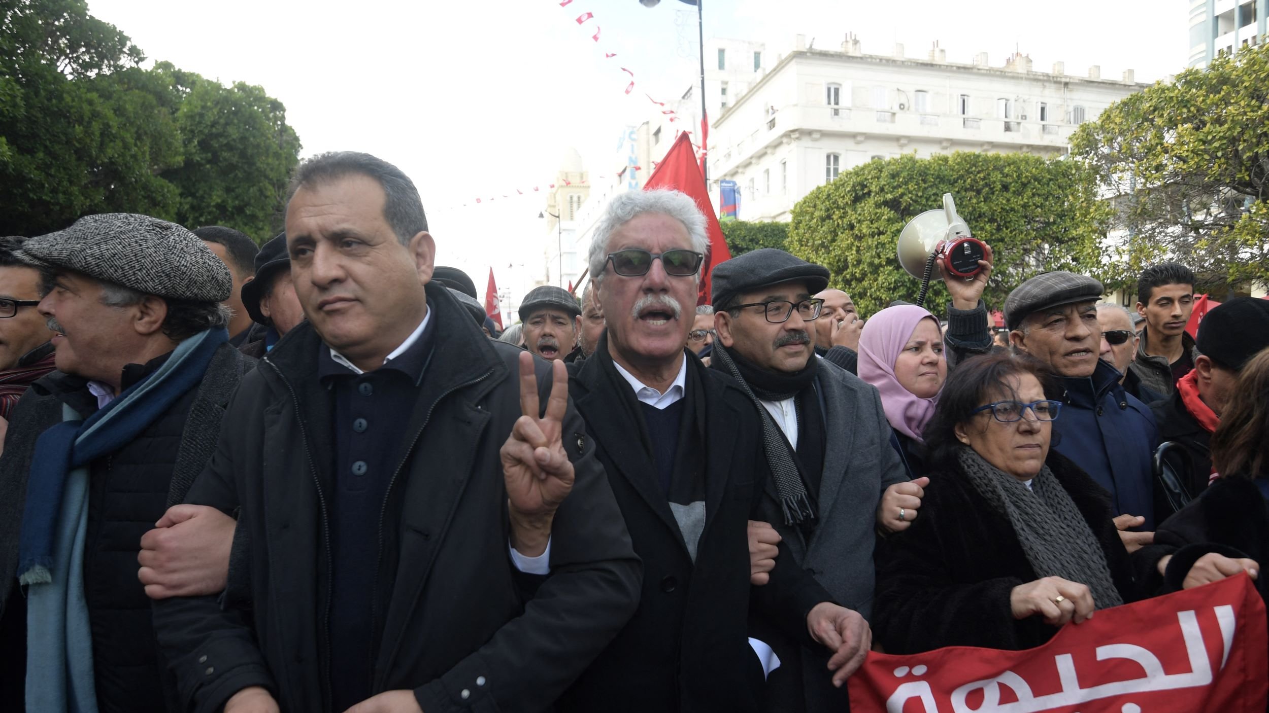 L'image montre un groupe de personnes rassemblées lors d'une manifestation. Au premier plan, on peut voir des hommes et des femmes exprimant des émotions vives, certains levant des signes de paix avec leurs mains. Les participants portent des vêtements variés, incluant des manteaux et des chapeaux. Des banderoles ou des drapeaux en arrière-plan ajoutent de la couleur à la scène, et l'atmosphère semble dynamique et engagée. Le décor environnant suggère un cadre urbain.
