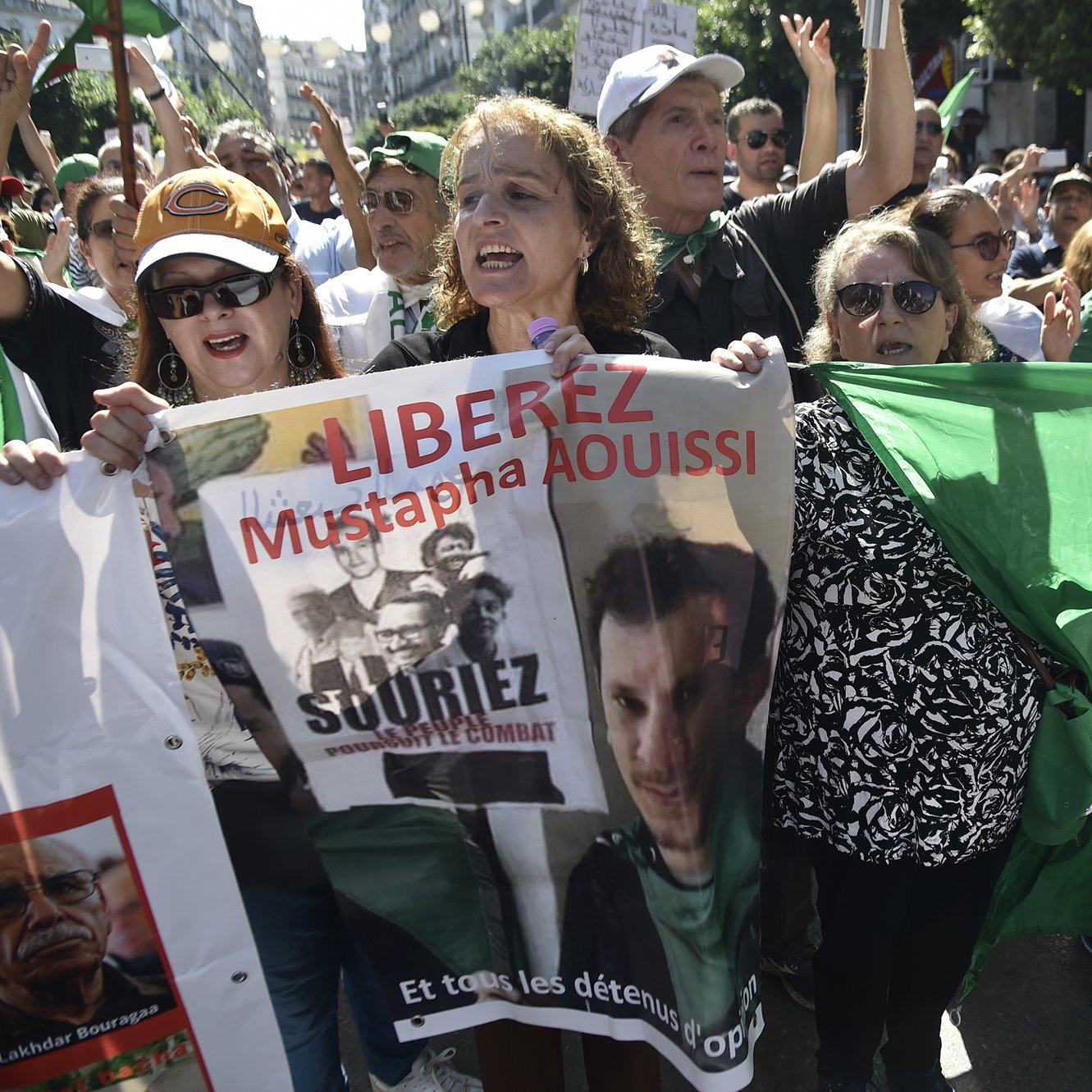 الصورة تظهر مجموعة من المتظاهرين في الجزائر، يحملون لافتات وأعلام. يبدو أن هناك نساء ورجال في المقدمة، ويعبرون عن مطالبات سياسية. إحدى اللافتات تحمل اسم "مصطفى عويصي" وعبارة "حرروا". الأجواء توحي بالحماسة والمطالبة بالتغيير.