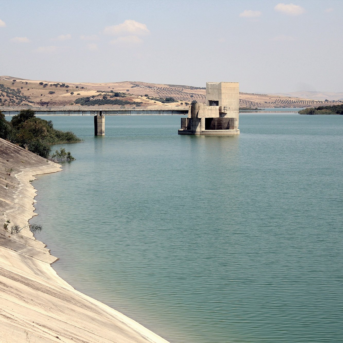 L'image montre un paysage calme avec un grand plan d'eau, probablement un réservoir ou un lac. Sur la gauche, la rive est recouverte d'une surface lisse et légèrement inclinée, tandis qu'à droite, on peut voir une structure en béton isolée au milieu de l'eau, avec des piliers qui émergent. À l'arrière-plan, des collines arrondies se dressent, ajoutant une touche de verdure au paysage. Le ciel est partiellement nuageux, ce qui crée une atmosphère paisible et naturelle.