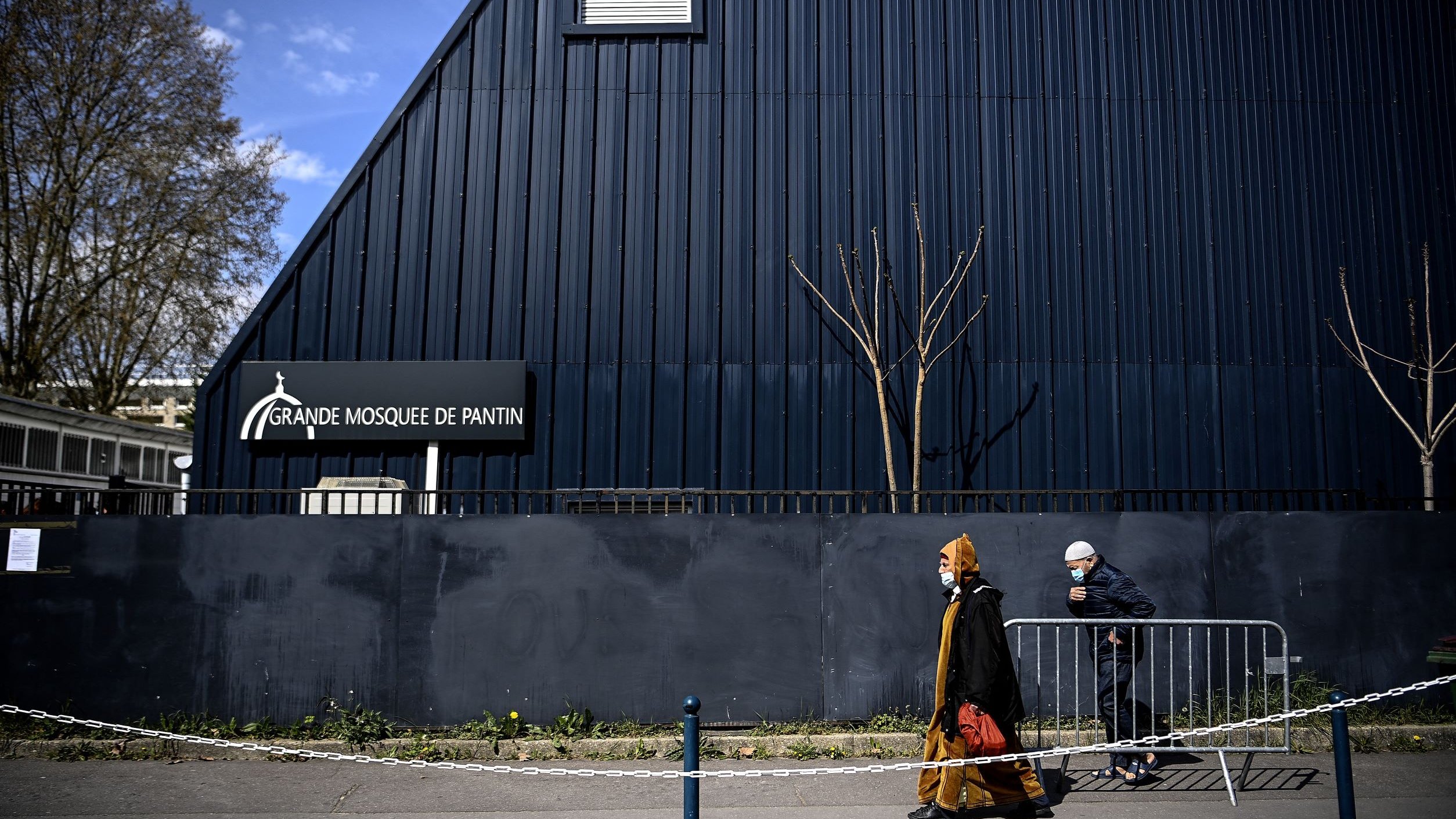L'image montre une scène extérieure près d'un bâtiment. On peut voir la façade d'une grande mosquée, identifiée par un panneau indiquant "Grande Mosquée de Pantin". À l'avant-plan, deux personnes marchent le long d'un chemin, l'une d'elles portant un voile et une tenue traditionnelle, tandis que l'autre est vêtue d'une manière plus décontractée, portant également un masque. L'architecture du bâtiment est moderne, avec un toit incliné, et l'environnement semble urbain, avec quelques arbres dépouillés de feuilles. Une barrière de sécurité sépare la rue du bâtiment.