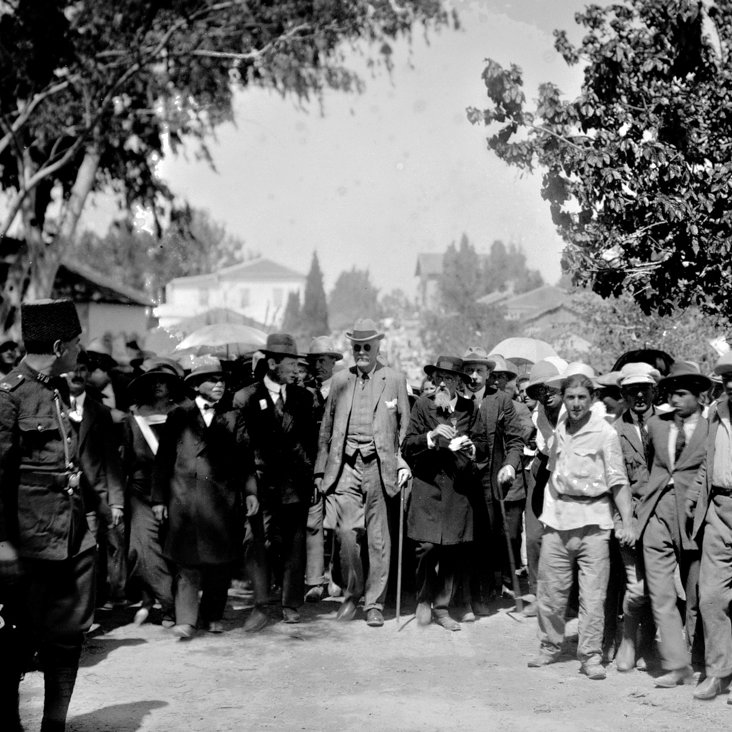 L'image montre un groupe de personnes rassemblées en extérieur, avec une scène animée. Au centre, une figure éminente se déplace, soutenue par une canne. De nombreuses personnes autour d'elle portent des chapeaux et tiennent des parasols, probablement pour se protéger du soleil. Des hommes en costume se distinguent parmi la foule, et l'environnement semble être urbain, avec des bâtiments en arrière-plan. La scène dégage une atmosphère d'importance ou de célébration.