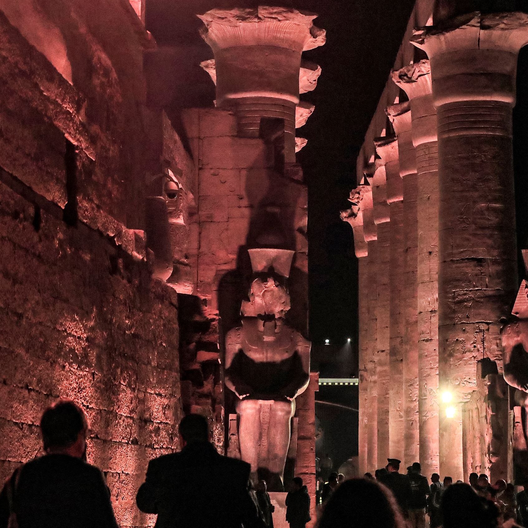 L'image présente un site historique, probablement des ruines égyptiennes, éclairées par des lumières tamisées, ce qui crée une atmosphère mystérieuse. On peut voir de grandes statues de pharaons ou de divinités, flanquant une structure impressionnante faite de colonnes. Les visiteurs se déplacent à travers le site, ajoutant une dimension humaine à cette scène ancienne, où l'architecture et les sculptures témoignent de la grandeur de la civilisation égyptienne. Les ombres et la lumière jouent un rôle important, accentuant les détails des sculptures et des murs.