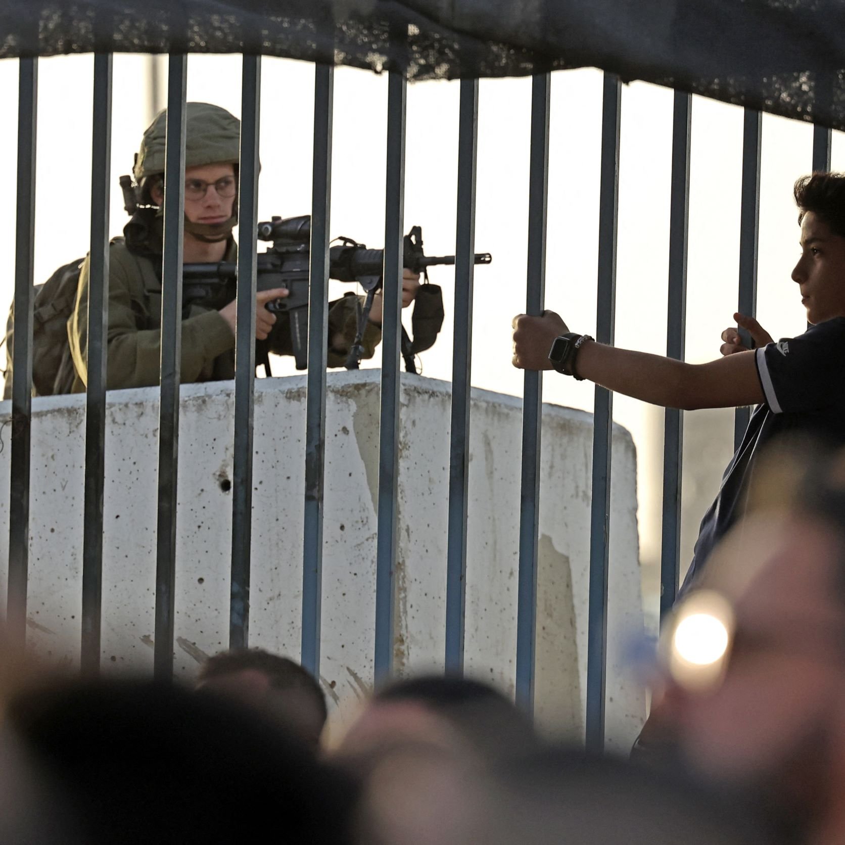 L'image montre un soldat armé stationné derrière une barrière ou un grillage. Il est en position de vigilance, regardant au loin. En face de lui, un jeune garçon semble gesticuler, tandis que d'autres enfants ou adolescents se trouvent à proximité. L'environnement semble tendu, reflétant une situation de contrôle ou de conflit. La lumière du soleil indique un moment de la journée où l'activité est encore présente.