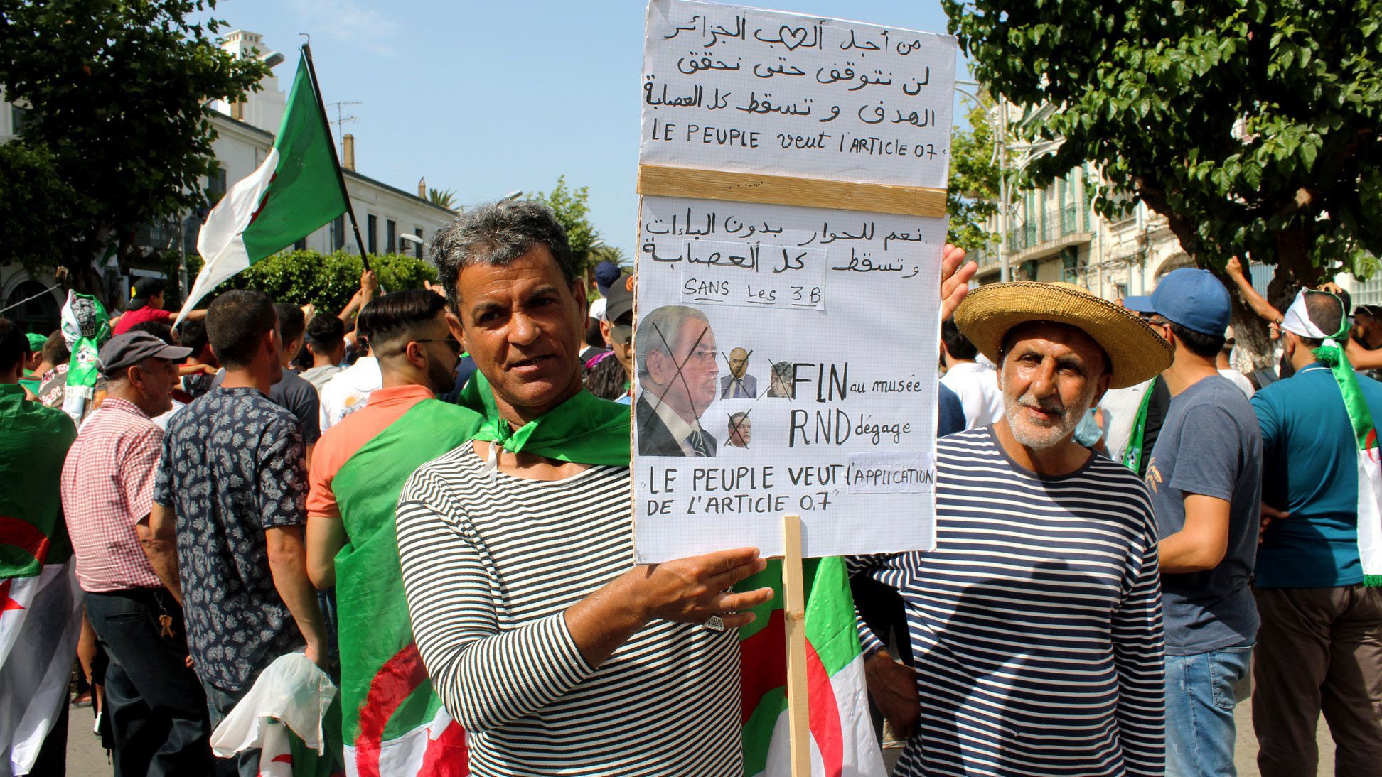 L'image montre un groupe de personnes manifestant, principalement des hommes, certains portant des drapeaux algériens. Deux manifestants se tiennent en avant-plan, l'un tenant une pancarte avec des inscriptions en arabe et en français. La pancarte semble exprimer des revendications politiques, notamment la fin de certaines pratiques liées à la politique algérienne. Les manifestants semblent unis et déterminés, se rassemblant pour faire entendre leur voix. L'atmosphère générale est celle d'une protestation pacifique.