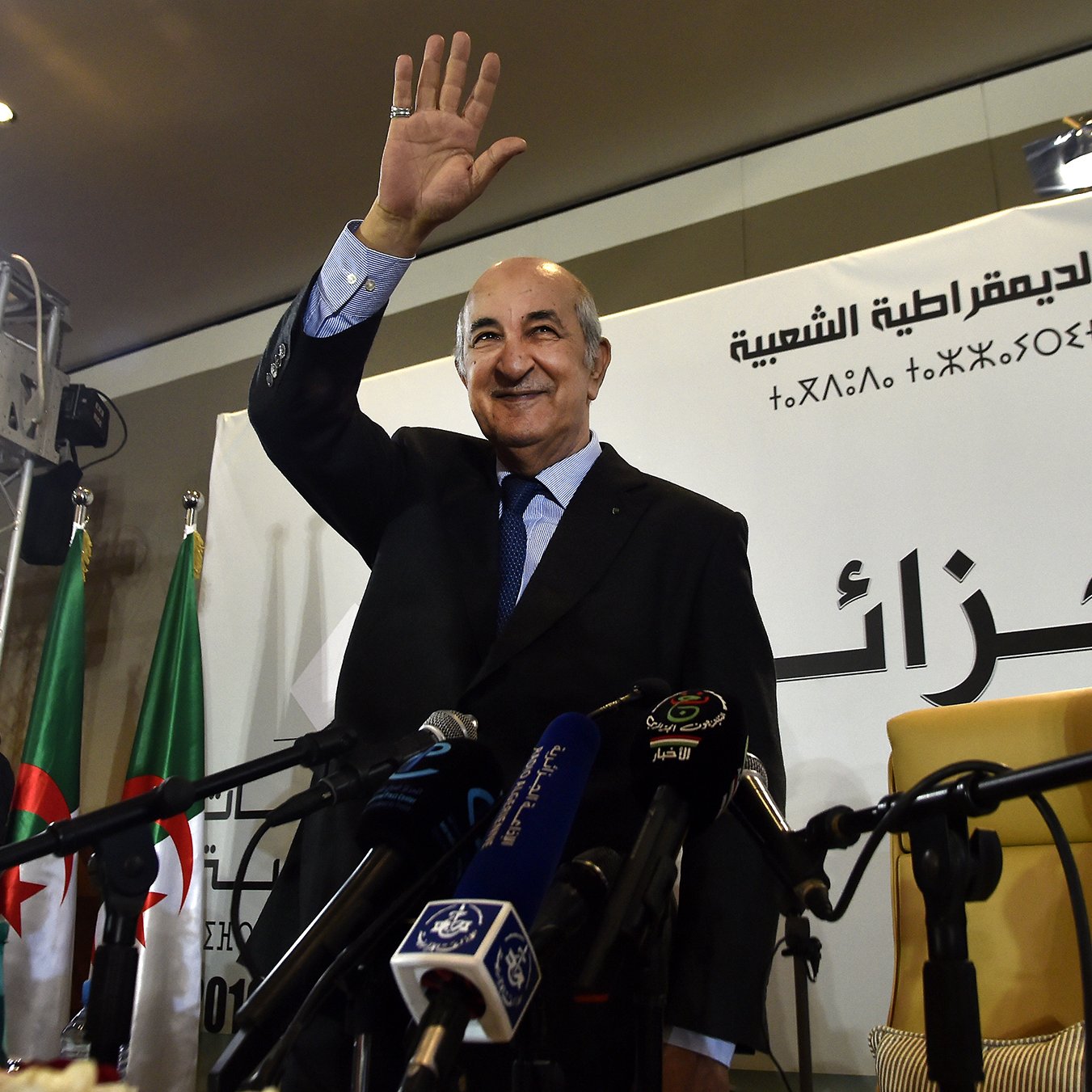 L'image montre un homme qui semble saluer une audience, debout devant un podium. Il porte un costume et a une main levée en signe de salut. En arrière-plan, on peut apercevoir des drapeaux, possiblement de l'Algérie, et des micros disposés devant lui, ce qui indique qu'il s'agit d'un événement officiel ou d'une conférence de presse. Des personnes semblent également présentes, apportant une ambiance formelle à la scène.
