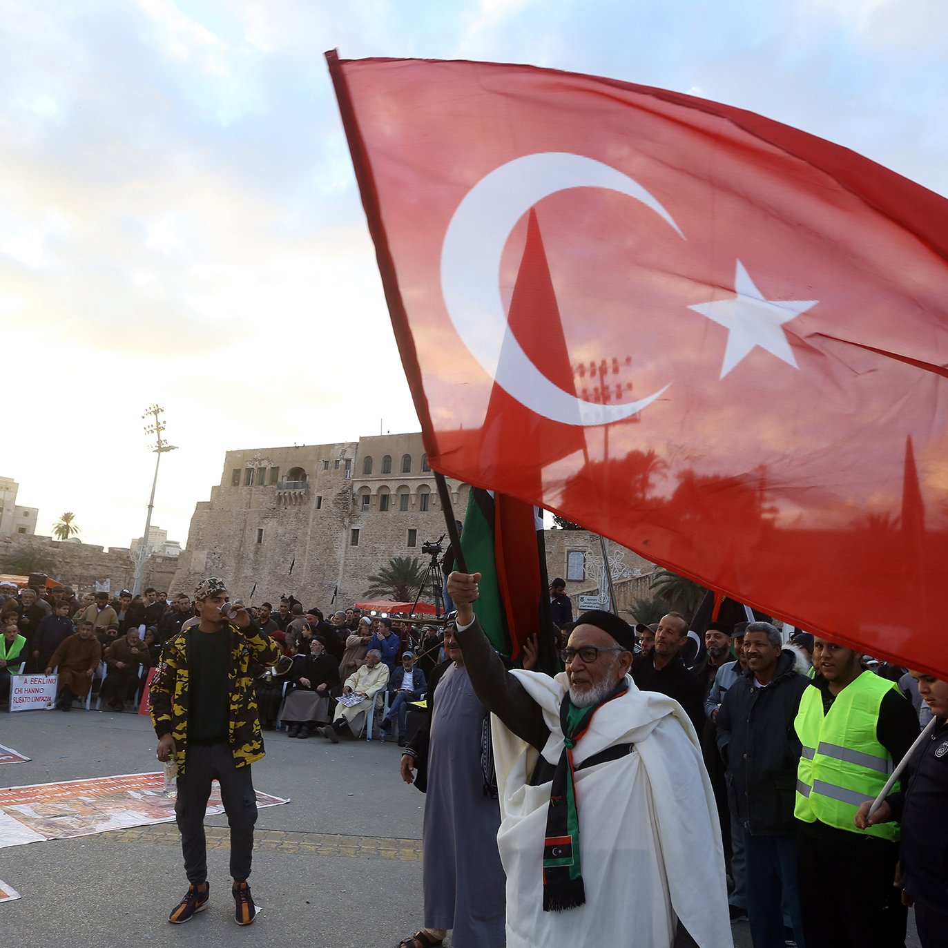 در این تصویر، جمعیتی در یک فضای باز مشاهده می‌شود که به نظر می‌رسد در حال برگزاری تجمعی هستند. یک مرد مسن با چهره‌ای شاد، پرچم بزرگ ترکیه را در دست دارد و به سوی دوربین اشاره می‌کند. در پس‌زمینه، ساختمان‌هایی قدیمی دیده می‌شود که نشان‌دهندهٔ تاریخ و فرهنگ خاصی هستند. مردم دیگری نیز در اطراف او به صورت جمعی ایستاده‌اند و تعدادی از آن‌ها لباس‌های زرد روشن پوشیده‌اند. آسمان ابری است و جلوه‌ای از نشاط و همبستگی در تصویر وجود دارد.