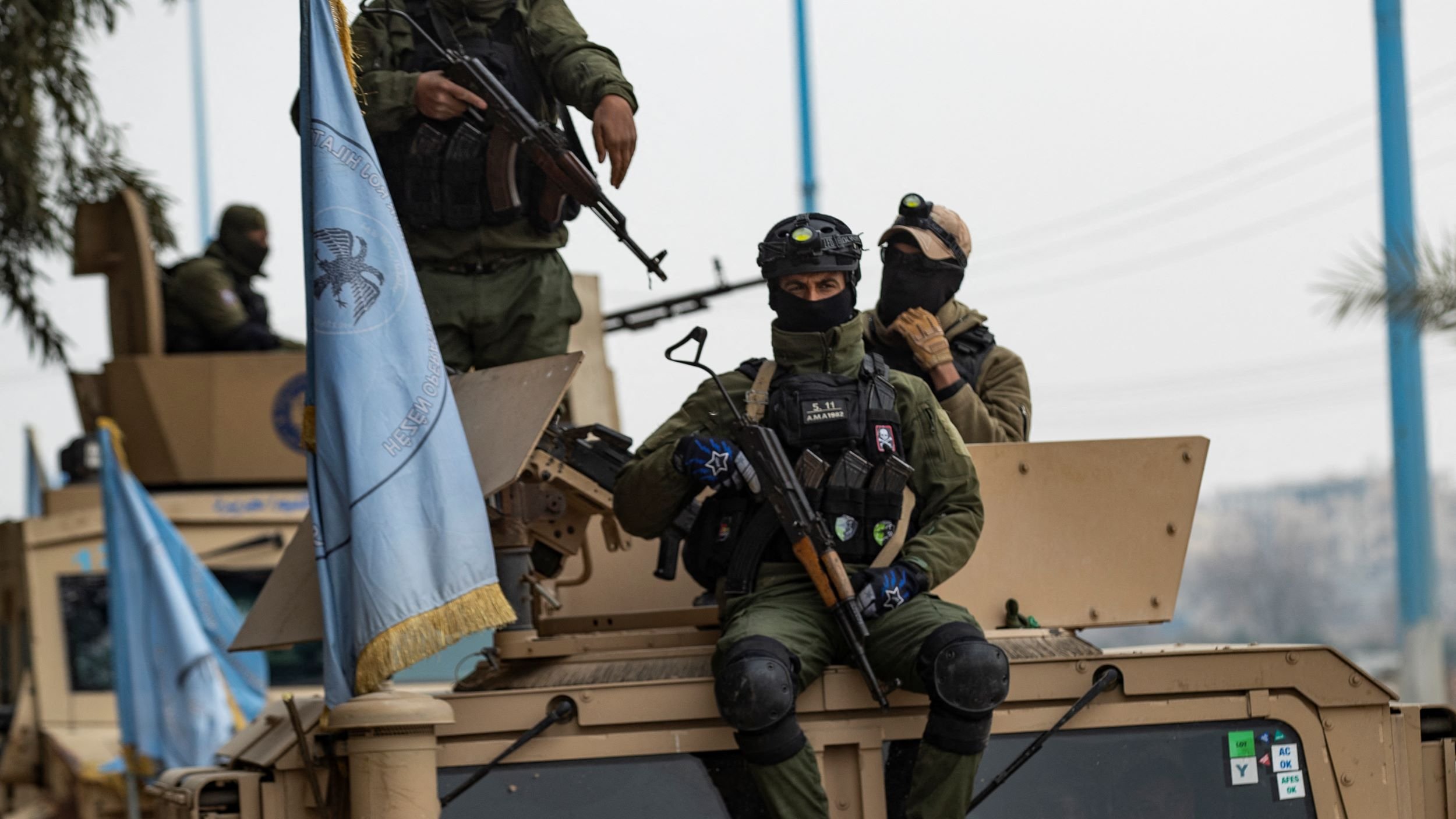 این تصویر گروهی از نیروهای مسلح را نمایش می‌دهد که روی یک وسیله نقلیه نظامی نشسته‌اند. نیروها لباس‌های نظامی تیره و کلاه‌خود دارند و برخی از آن‌ها سلاح‌های جداگانه‌ای در دست دارند. بر روی وسیله نقلیه پرچمی و نمادهای مربوط به سازمان‌های بین‌المللی یا نظامی به چشم می‌خورد. پس‌زمینه تصویر به وضوح نشان‌دهنده یک محیط خارج از شهر است.