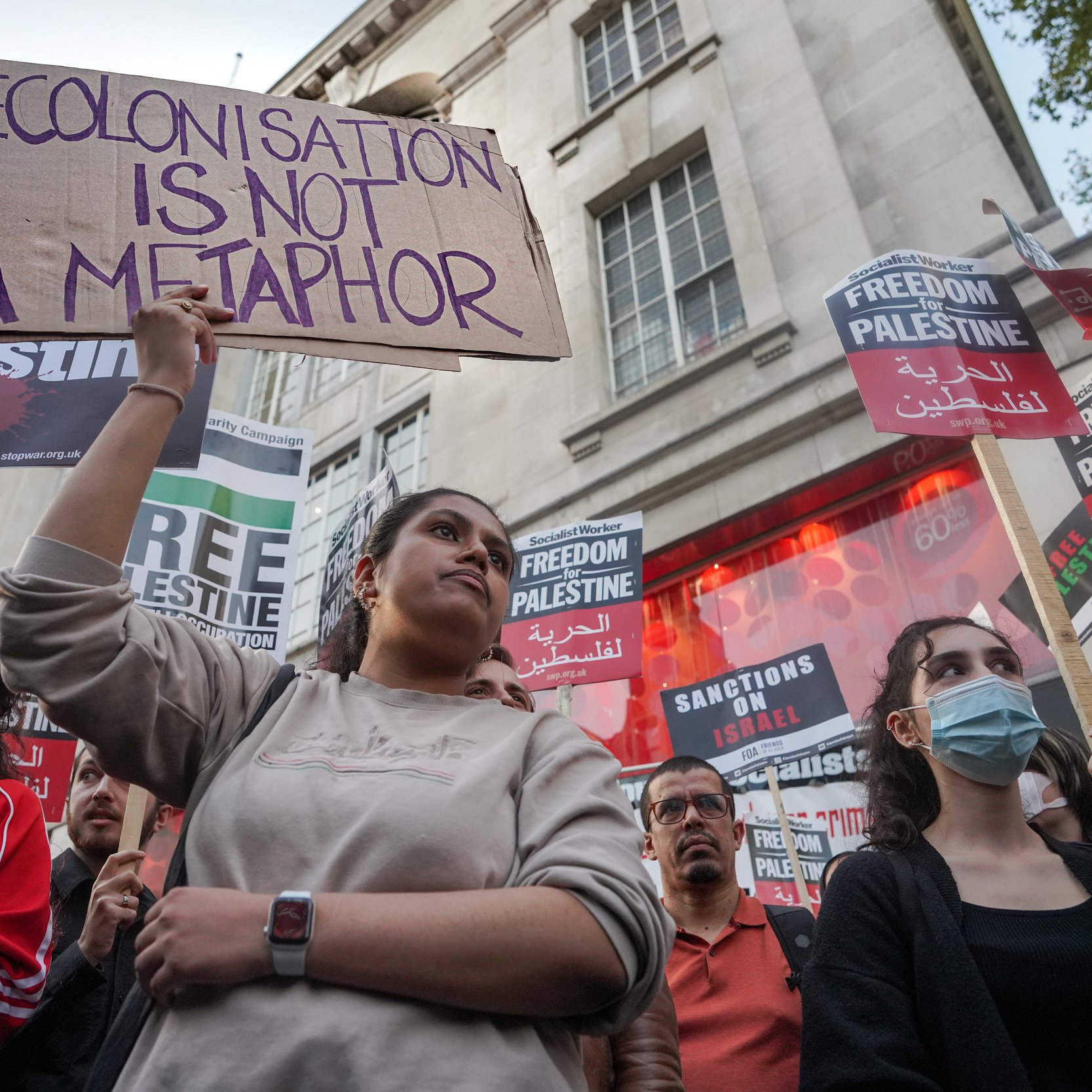 L'immagine mostra un gruppo di persone in una manifestazione, molte delle quali tengono in mano cartelli con frasi di protesta. Una giovane donna al centro tiene un cartello che dice "La decolonizzazione non è una metafora". Sullo sfondo, ci sono altri cartelli con scritte come "Libertà per la Palestina" e "Sanzioni a Israele". I partecipanti sembrano esprimere il loro sostegno per la causa palestinese, mentre l'atmosfera è di attivismo e impegno sociale.