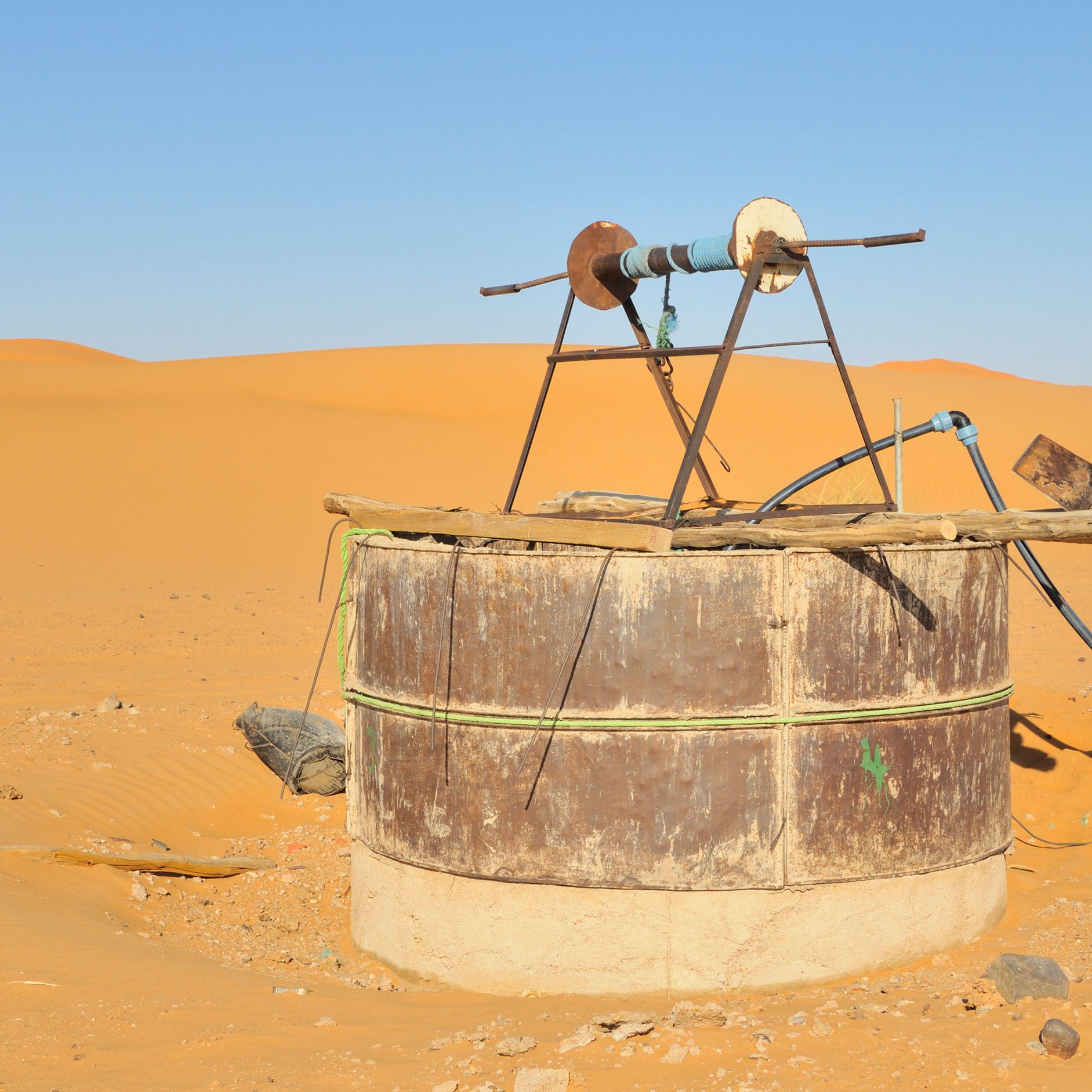 L'image montre une structure située dans un désert de sable doré. Au centre, il y a un puits ou un réservoir en métal, recouvert de tôle, avec un mécanisme en bois au-dessus. Ce dernier semble être utilisé pour pomper de l'eau. La terre environnante est sèche et aride, et on aperçoit des dunes de sable qui s'étendent à perte de vue sous un ciel clair et sans nuages. L'ensemble évoque un environnement désertique, isolé et dépouillé.