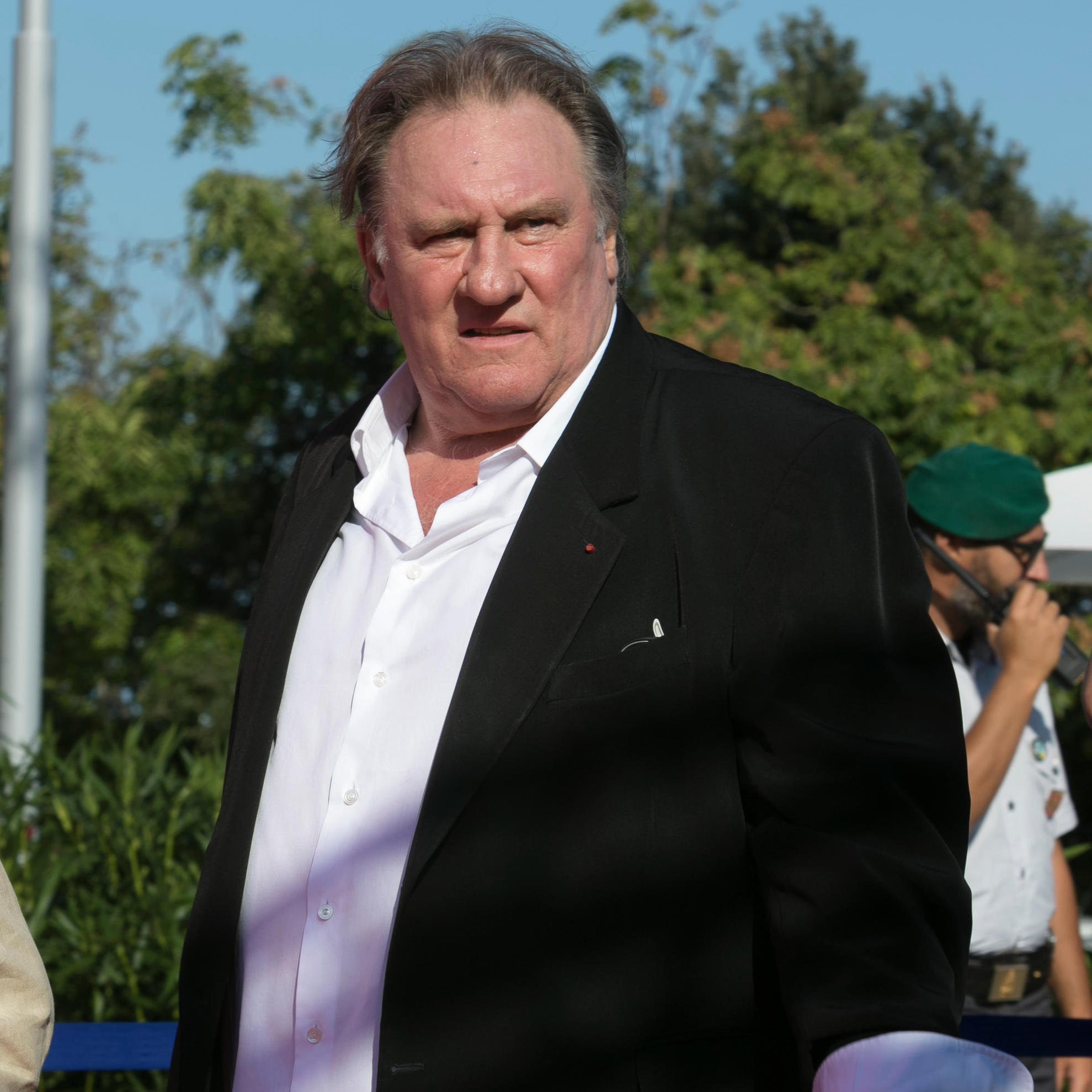 Je ne sais pas qui apparaît sur cette image. Cependant, je peux décrire certains éléments visuels. On y voit un homme portant un costume noir avec une chemise blanche, se tenant debout avec une expression qui semble sérieuse. L'environnement présente des arbres et une ambiance ensoleillée. Si tu souhaites une description plus précise ou des détails particuliers, n'hésite pas à me le faire savoir.