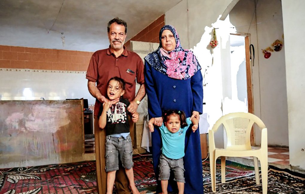 L'image montre une famille pose ensemble dans une pièce chaleureuse. Un homme et une femme, avec deux enfants, se tiennent debout sur un tapis. La femme porte un hijab et une longue robe, tandis que les enfants semblent joyeux. En arrière-plan, on peut voir des murs et une cuisine simple. L'atmosphère semble conviviale et familiale.