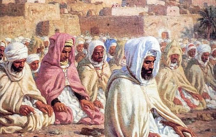 L'image représente un groupe de personnes rassemblées en prière, probablement dans un contexte religieux. Elles sont vêtues de robes traditionnelles et portent des turbans ou des voiles. Les hommes semblent concentrés et méditatifs, assis sur le sol. En arrière-plan, on peut voir une ville avec des maisons aux couleurs claires, sous un ciel bleu, ce qui suggère un environnement désertique ou méditerranéen. L'atmosphère est paisible et spirituelle.