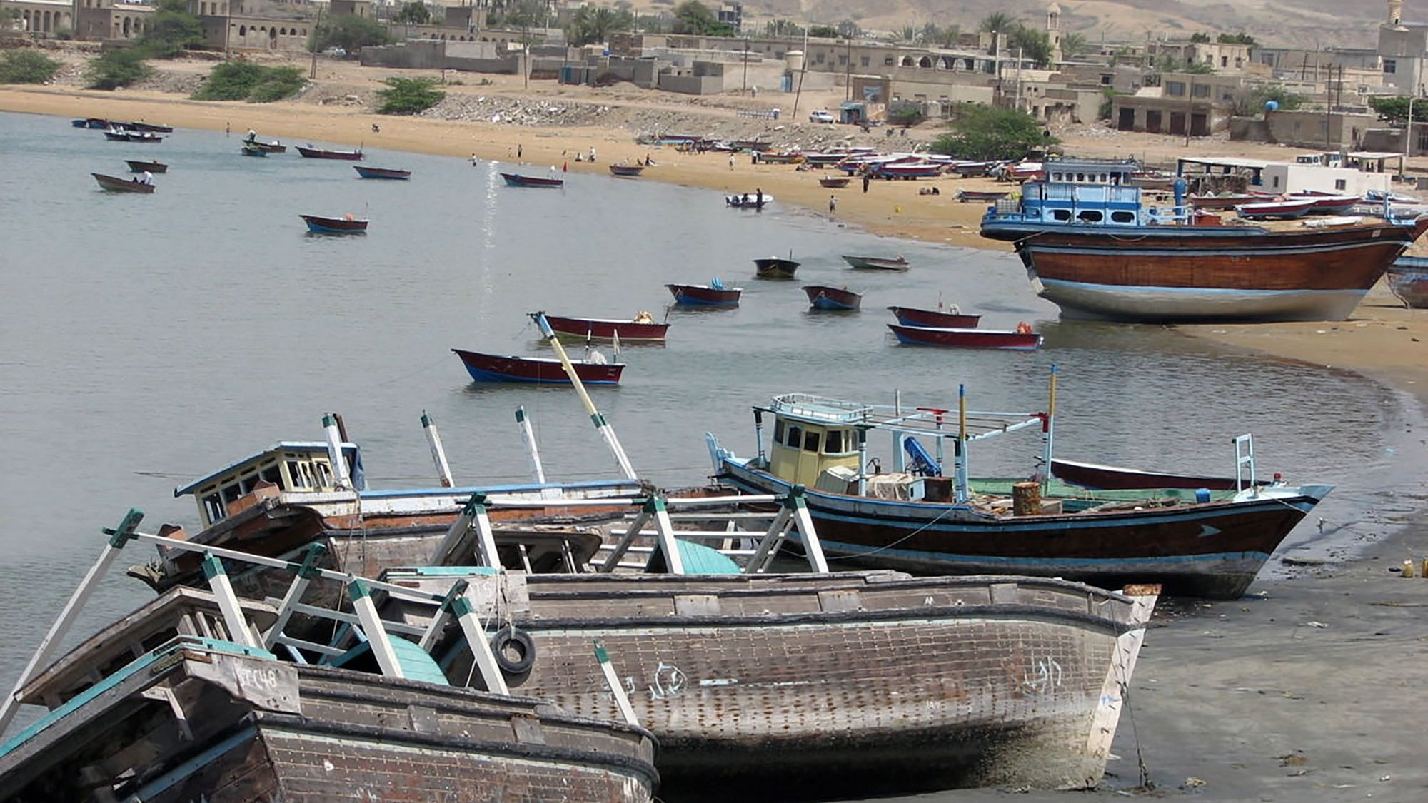 L'image montre une scène côtière avec plusieurs bateaux de pêche échoués sur la plage. On peut voir des petites embarcations peintes en différentes couleurs, certaines abîmées, qui sont disposées le long d'une côte sablonneuse. En arrière-plan, des bâtiments témoignent d'une ville ou d'un village, et le paysage est dominé par des collines ou des falaises. L'eau est calme, et l'atmosphère semble tranquille, avec quelques personnes visibles sur la plage.
