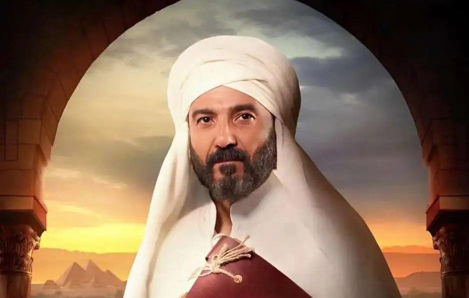 L'image représente un homme portant un turban et une tenue traditionnelle. Il a une barbe et une expression sérieuse. En arrière-plan, on aperçoit un paysage désertique avec des pyramides, suggérant une ambiance historique ou culturelle. La lumière semble douce, ajoutant une atmosphère dramatique à la scène.