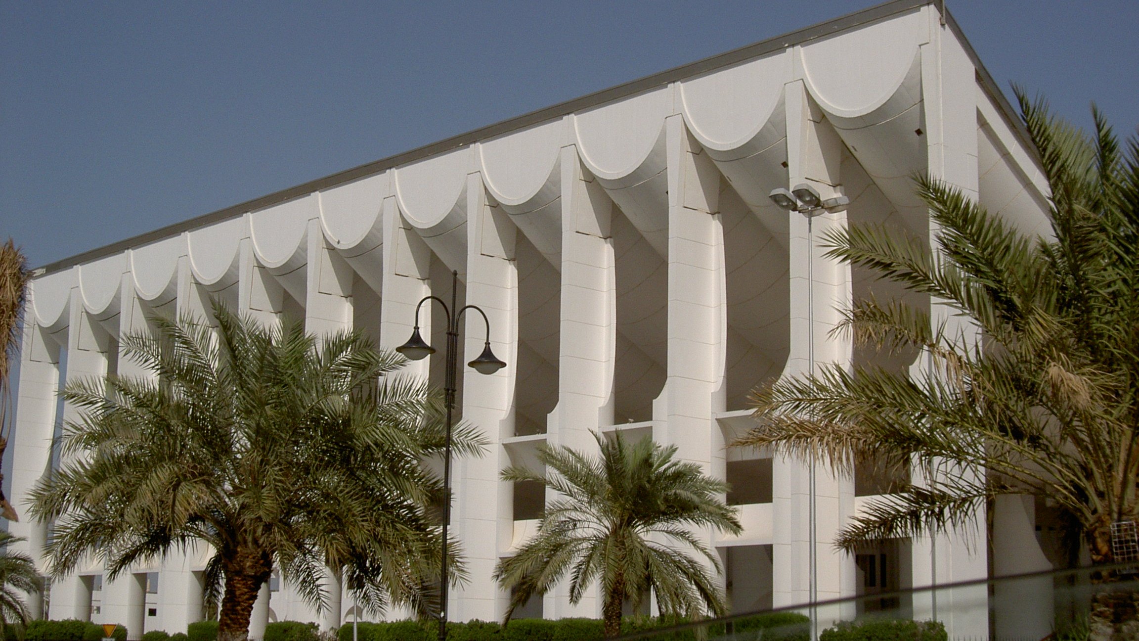 L'image montre un bâtiment moderne avec une architecture distinctive. La façade est principalement blanche, ornée de structures en forme de vagues ou de voiles sur le haut. Des colonnes soutiennent la structure, ajoutant une touche de sophistication. On peut également observer des palmiers plantés devant le bâtiment, ce qui donne une atmosphère tropicale et accueillante. Le ciel est clair et bleu, accentuant la luminosité de l'ensemble.