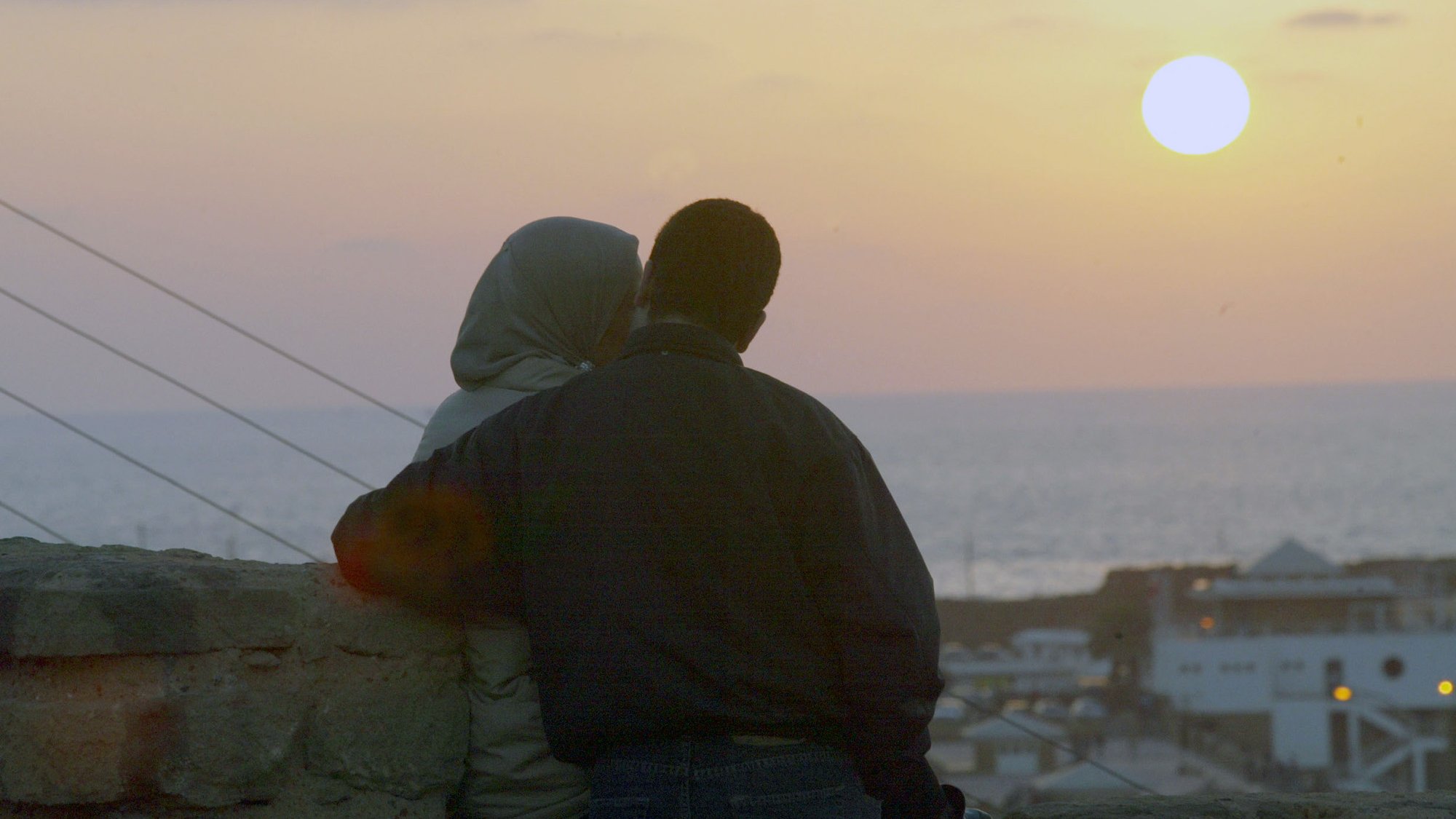 L'image montre un couple assis côte à côte, observant le coucher de soleil. L'un des membres du couple porte un hijab, tandis que l'autre est vêtu d'une veste sombre. Ils semblent se rapprocher l'un de l'autre, créant une atmosphère intime. En arrière-plan, on peut voir la mer, et le ciel est teinté de couleurs douces, avec le soleil se couchant à l'horizon. Les bâtiments sont légèrement visibles en bas, ajoutant à la scène romantique.