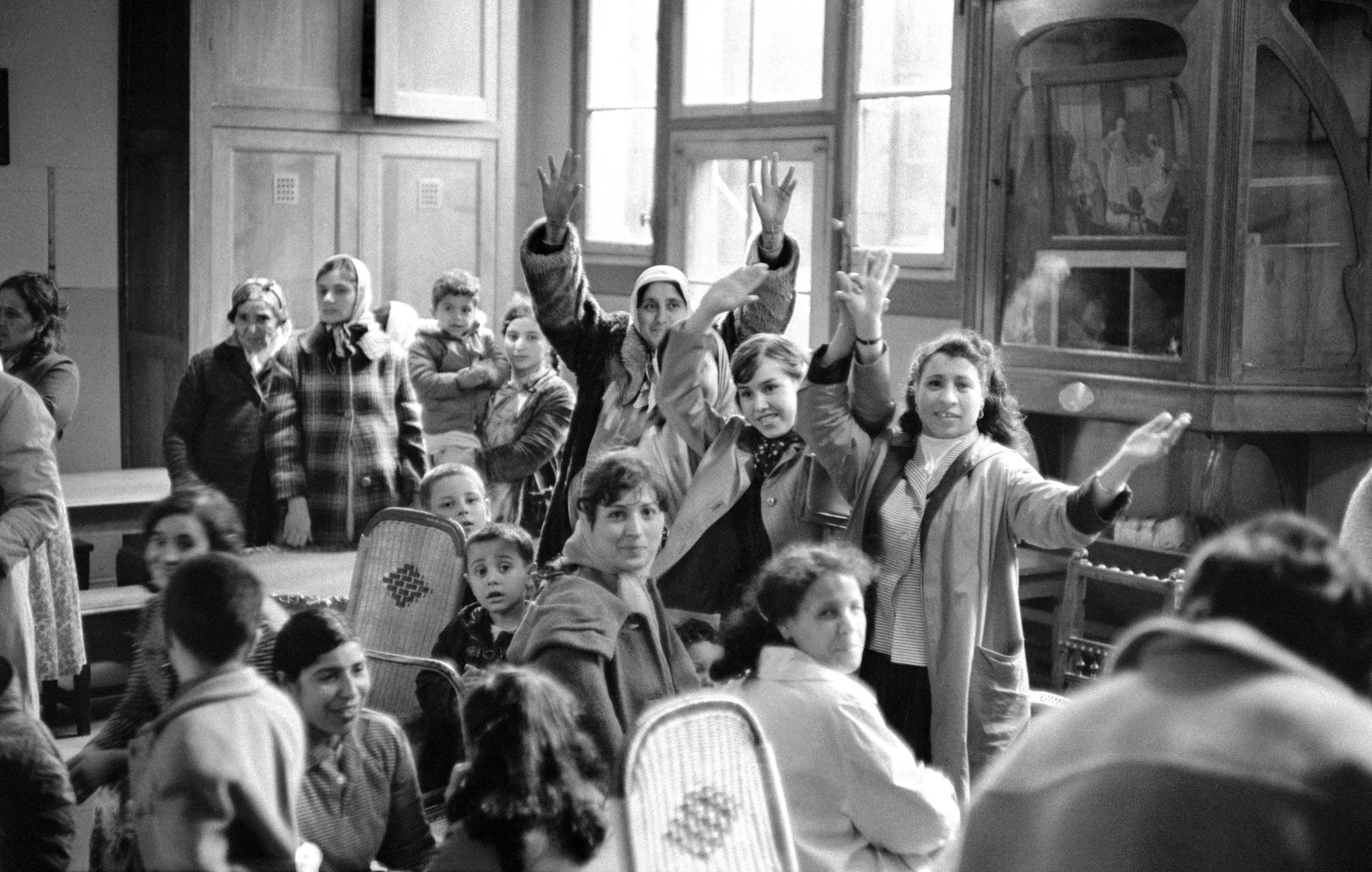 L'image en noir et blanc montre un groupe de personnes dans une pièce, probablement un lieu communautaire. Plusieurs adultes et enfants sont présents, certains levant les bras en signe d'encouragement ou d'émerveillement. L'atmosphère semble conviviale, avec des visages qui expriment de la joie ou de l'excitation. On peut voir des fauteuils en osier et un grand meuble en bois à l'arrière-plan, ajoutant une touche de chaleur à l'environnement. La lumière naturelle entre par de grandes fenêtres, illuminant la scène.