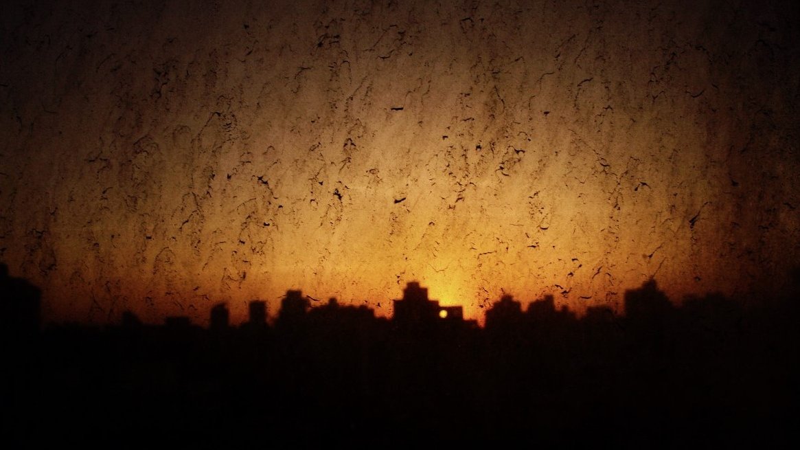 L'image montre un paysage urbain au crépuscule, avec des silhouettes de bâtiments se découpant contre un ciel orangé. La texture de la vitre est visible, ajoutant une dimension bruitée et floue à la scène. Les reflets et les imperfections de la vitre créent une atmosphère légèrement mystérieuse, tandis que la lumière du soleil couchant rappelle la fin de la journée.