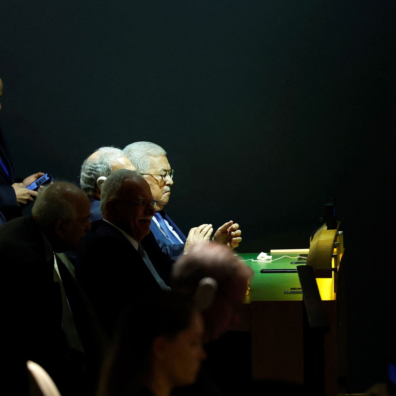 L'immagine mostra un gruppo di uomini seduti a un tavolo, apparentemente impegnati in una discussione o in un evento ufficiale. Alcuni di loro sembrano concentrati, mentre uno di loro sta guardando il cellulare. L'illuminazione è tenue, con un contrasto tra le zone illuminate e quelle in ombra, conferendo un'atmosfera seria e formale all'ambiente. Non ci sono elementi di identificazione chiara per i volti dei partecipanti.