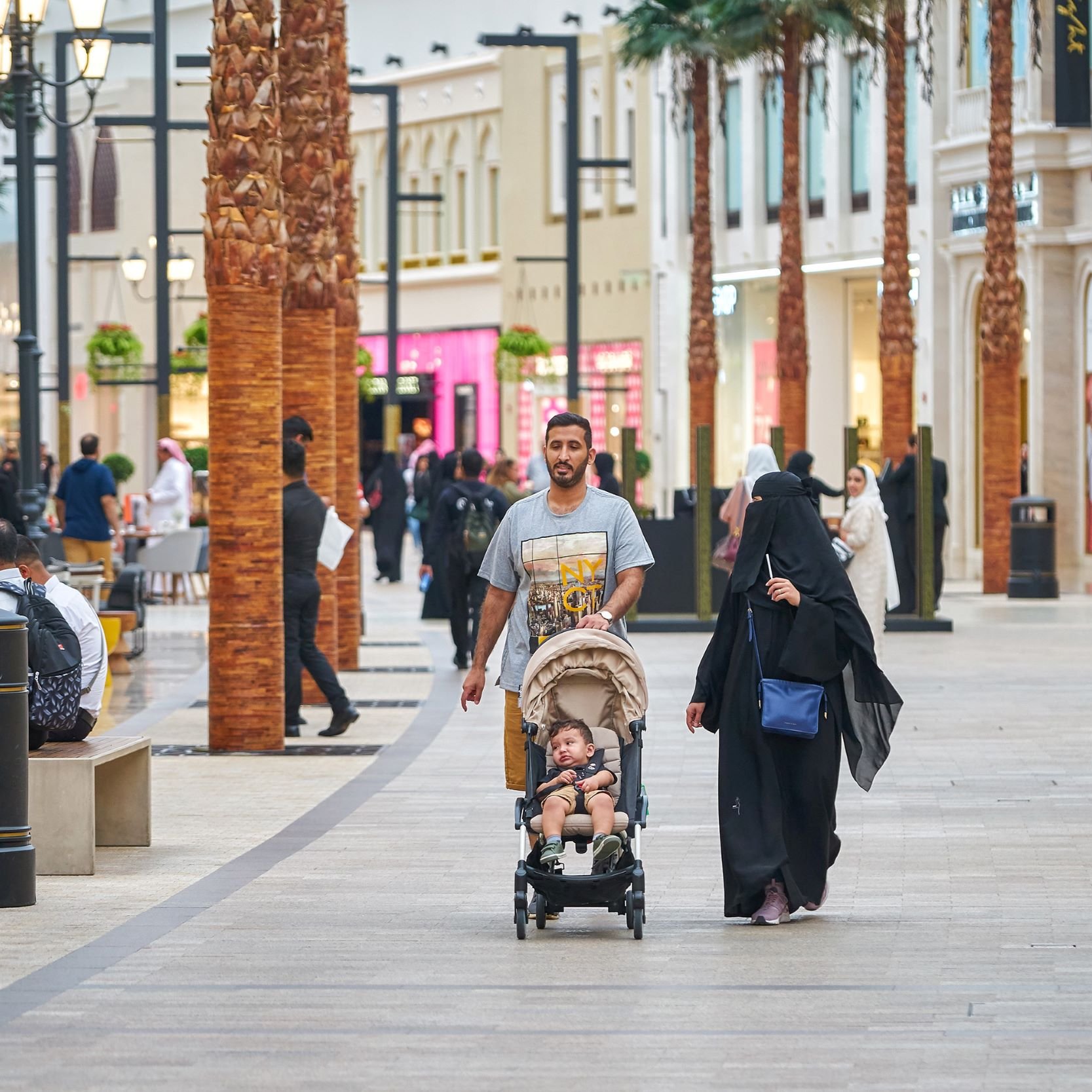 L'image montre une scène animée dans un centre commercial. On peut voir des palmés en arrière-plan et des magasins de différentes boutiques. Un homme marche avec un enfant dans une poussette, tandis qu'une femme, vêtue d'une abaya, les accompagne. Plusieurs autres personnes sont visibles dans l'allée, créant une ambiance de vie urbaine dynamique. La lumière est claire et l'atmosphère semble accueillante.