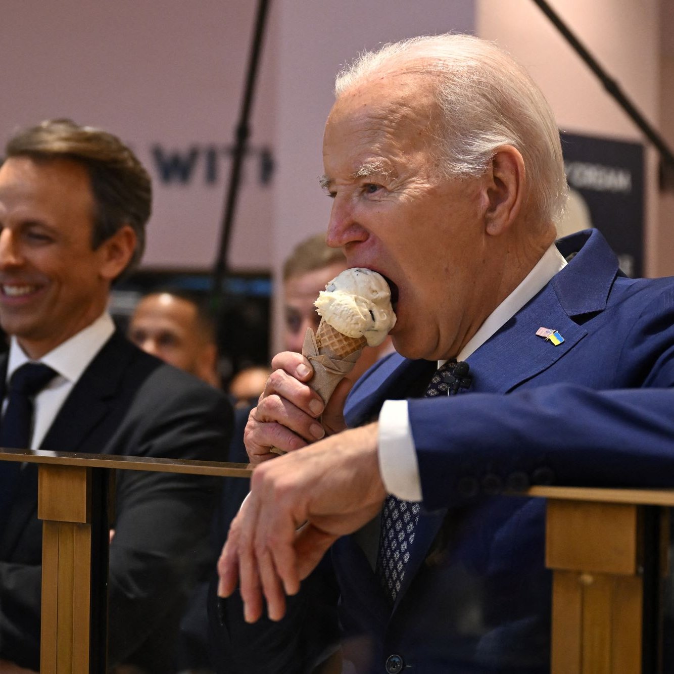 الصورة تُظهر رجلًا في بدلة رسمية وهو يأخذ قضمة من مثلجات، بينما يقف بجانبه رجل آخر يبتسم. يبدو أن المكان عبارة عن حدث أو اجتماع، حيث يوجد العديد من الأشخاص في الخلفية. الرجل الذي يأكل المثلجات هو شخص بارز يبدو سعيدًا وهو يستمتع باللحظة.