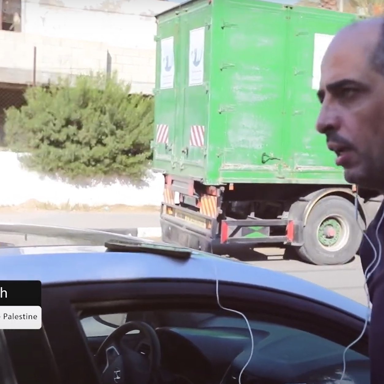 L'image présente un homme marchant à côté d'une voiture, portant des écouteurs. Il a un air concentré et semble être dans un environnement urbain avec un camion vert en arrière-plan. Un texte en bas de l'image mentionne "Bilal Jadallah, Directeur de Press House - Palestine".