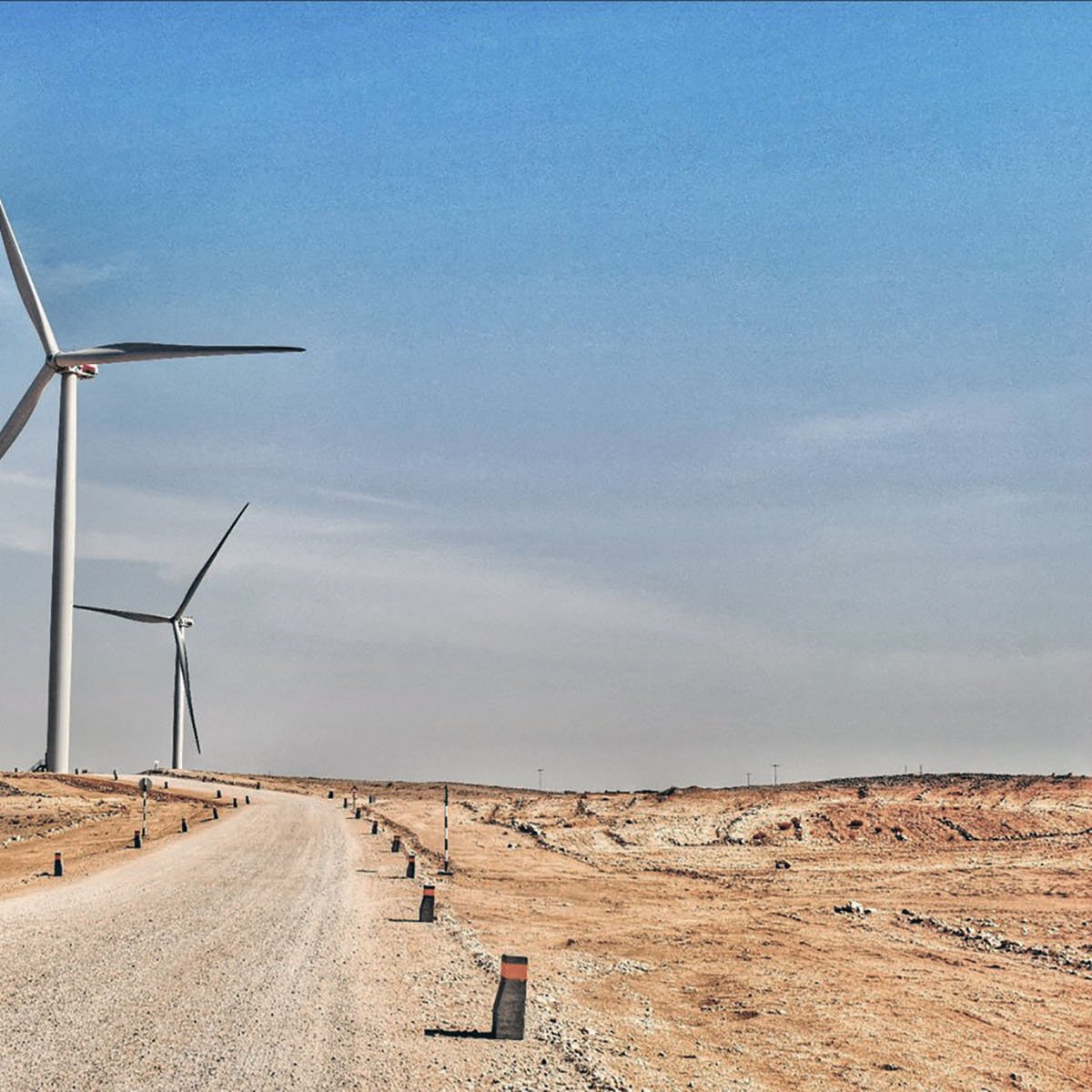 The image depicts a wide, unpaved road extending through a dry, barren landscape. On either side of the road, there are several large wind turbines standing tall against a clear blue sky. The terrain appears arid, with sparse vegetation and a sandy or rocky surface. The road is lined with small posts or markers, adding a structured element to the otherwise natural setting. The overall atmosphere conveys a sense of open space and renewable energy in a desert-like environment.